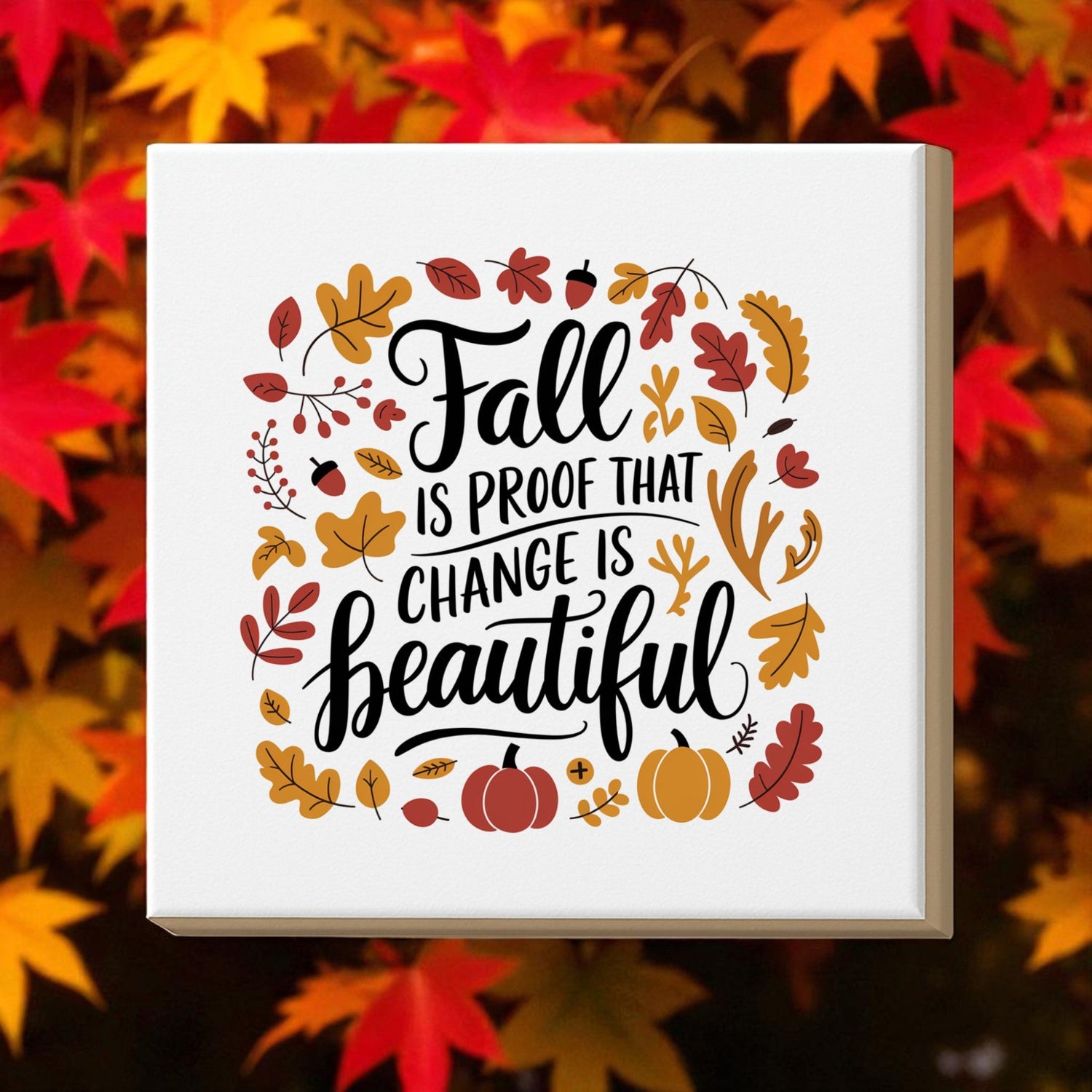 A decorative sign with autumn leaves and pumpkins reads, “Fall is proof that change is beautiful.” Bright orange, red, and yellow leaves are blurred in the background.