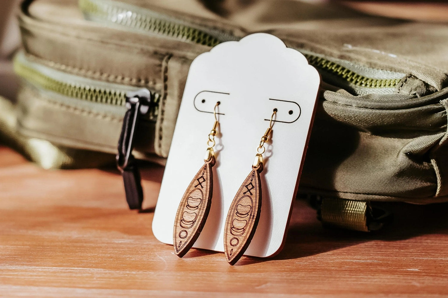 A pair of brown, canoe-shaped wooden earrings with gold hooks displayed on a white card, propped against a green backpack on a wooden surface.