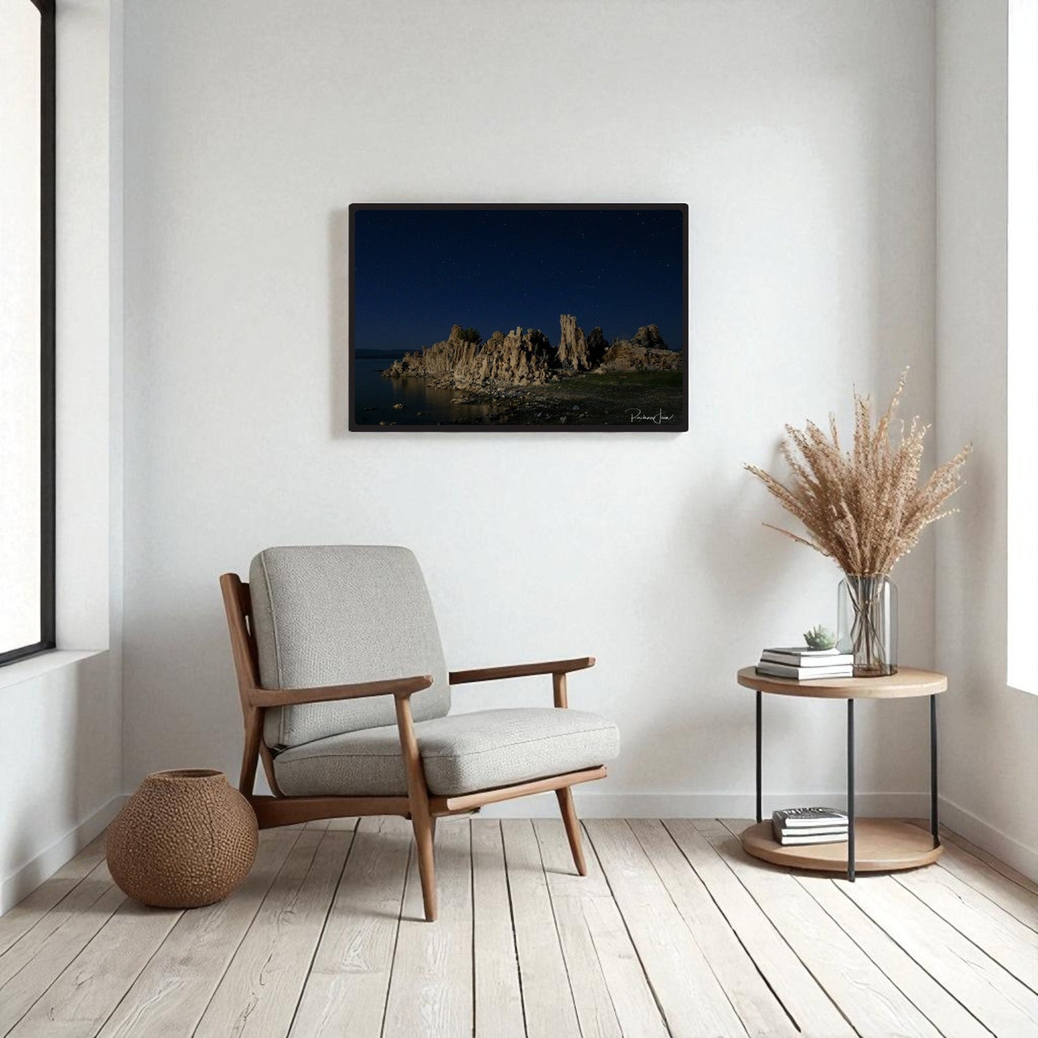 Minimalist living room with a light gray armchair, round side table with dried pampas grass, woven basket, and a landscape photograph of rock formations hanging on the wall.