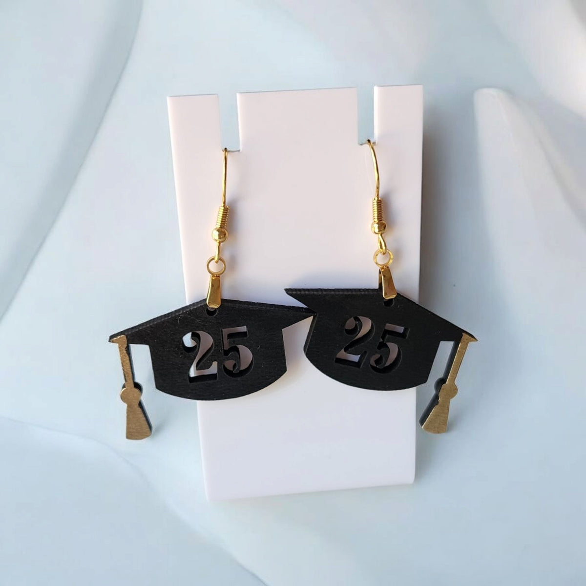Mirabilia Boutique Graduation Cap Earrings Class of 2025 feature black caps with gold tassels and a “25” cut-out, laser cut for a unique grad jewelry gift or party accessory. Displayed on a white stand; perfect for celebrating her big day!.