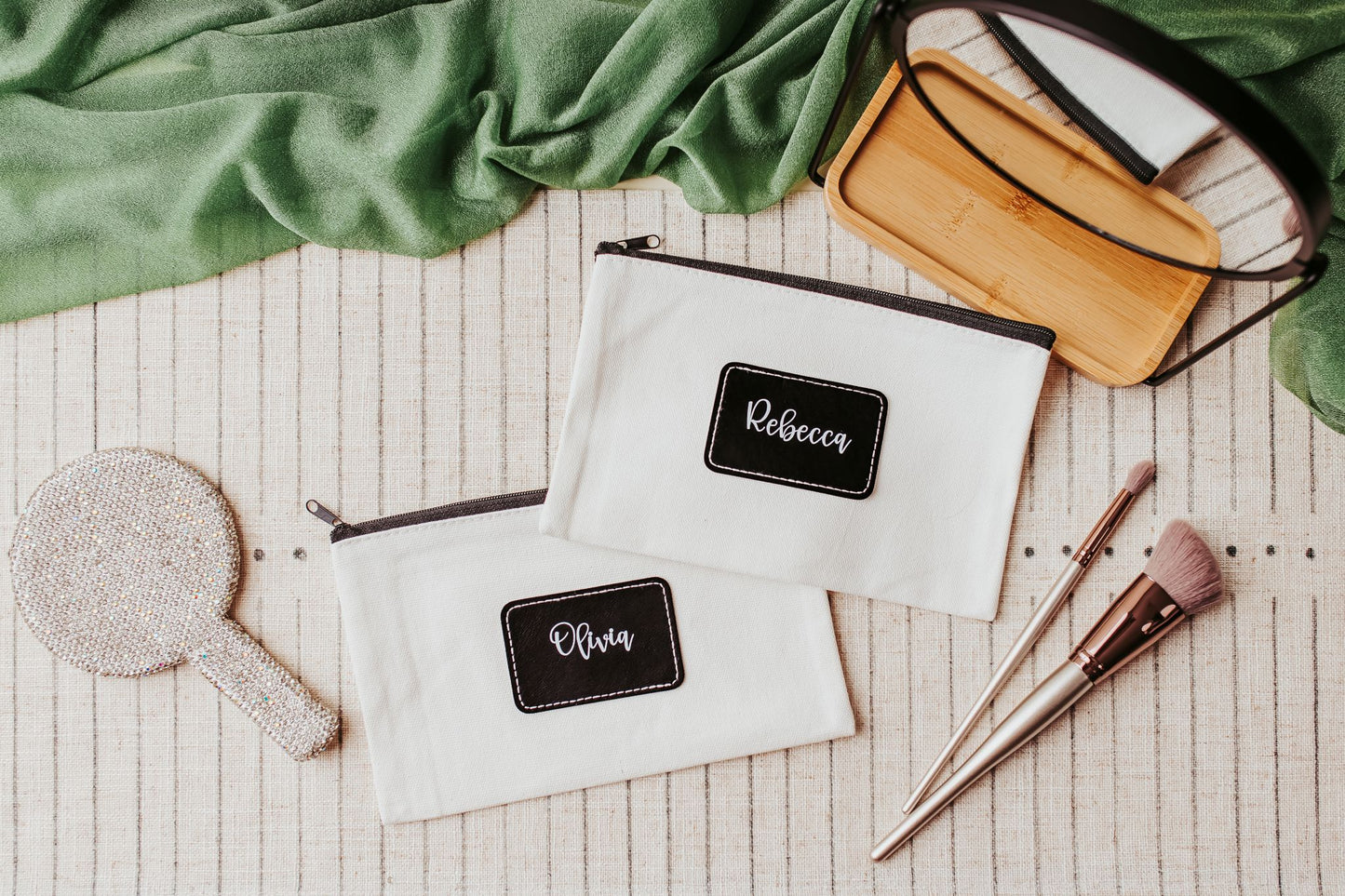 Two white Mirabilia Boutique Custom Name Makeup Bags, labeled “Rebecca” and “Olivia,” rest near beauty tools—a stylish bridesmaid gift or personalized canvas pouch from the Personalized Canvas Pouch collection.