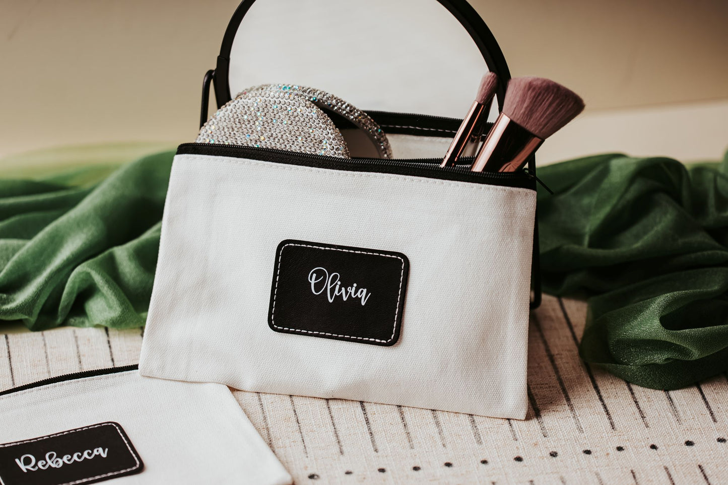 A Mirabilia Boutique Custom Name Makeup Bag labeled "Olivia" holds brushes and pads, while another personalized canvas pouch marked "Rebecca" rests nearby—both ideal bridesmaid gifts on a striped surface with green fabric in the background.