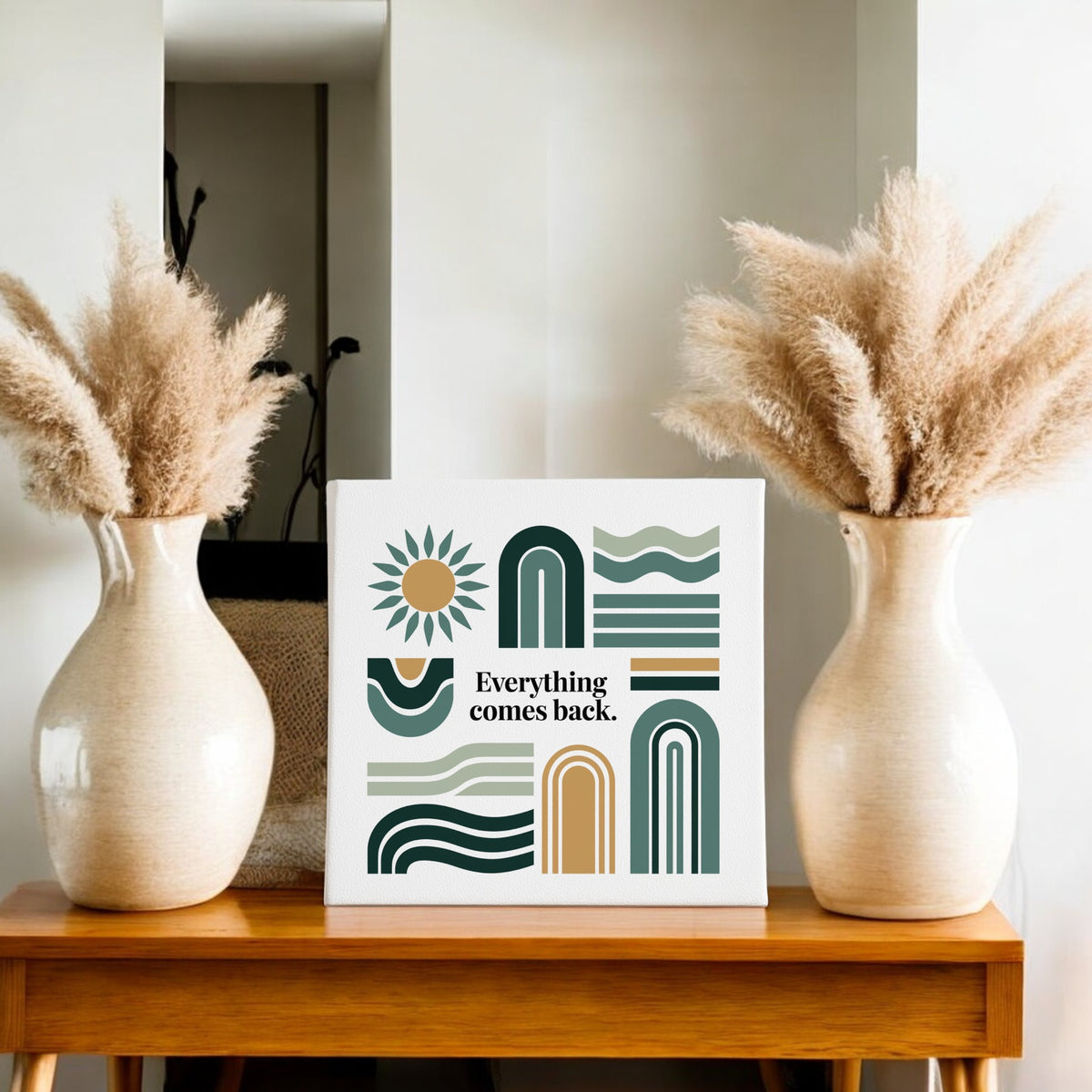 A wooden table displays two beige vases with pampas grass and the Mirabilia Boutique “Everything Comes Back Canvas – Earth Tone Wall Art,” featuring geometric shapes and earthy hues for modern decor.