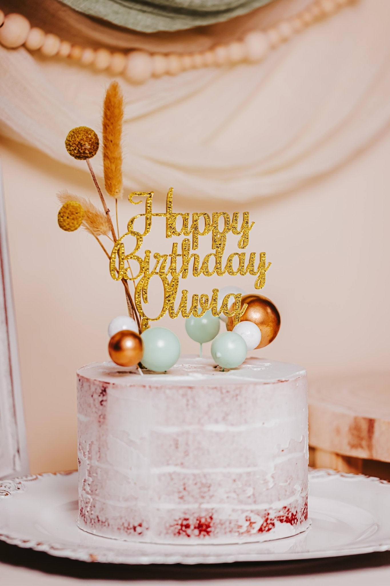 A lightly frosted cake featuring a golden Acrylic Happy Birthday Cake Topper from Mirabilia Boutique, personalized with "Happy Birthday Olivia," adorned with custom color accents and decorative elements such as spherical ornaments and dried flowers, elegantly presented against a soft, draped fabric backdrop.