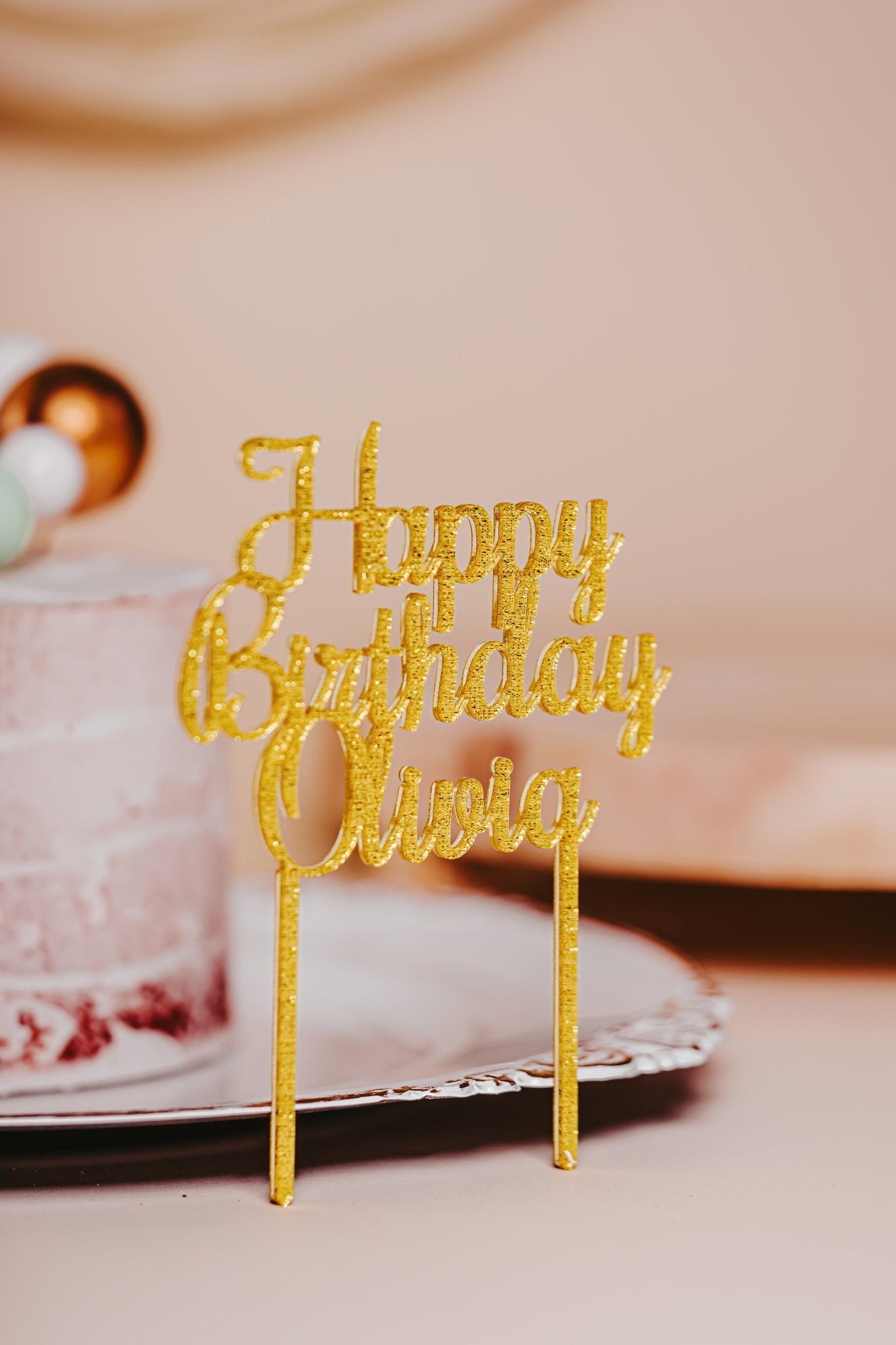 A stylish, glittery gold cake topper from Mirabilia Boutique, featuring the text "Happy Birthday Olivia," elegantly adorns a partially visible white cake with pink frosting. The softly blurred background enhances the prominence of this Acrylic Happy Birthday Cake Topper for Festive Celebrations, making it a perfect choice for any special occasion.