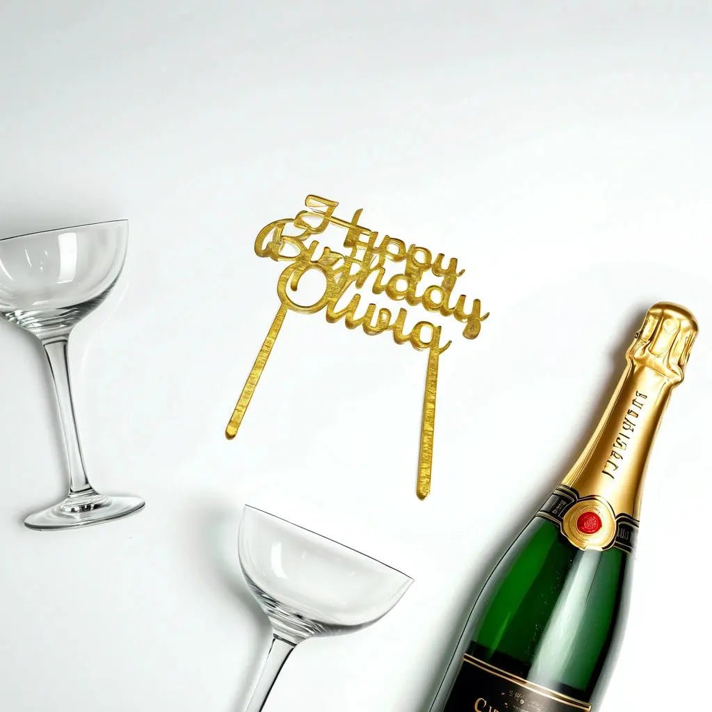 Two empty champagne glasses, a bottle of champagne, and the Mirabilia Boutique Acrylic Happy Birthday Cake Topper—featuring the elegant message "Happy Birthday Olivia"—are elegantly arranged on a pristine white background.