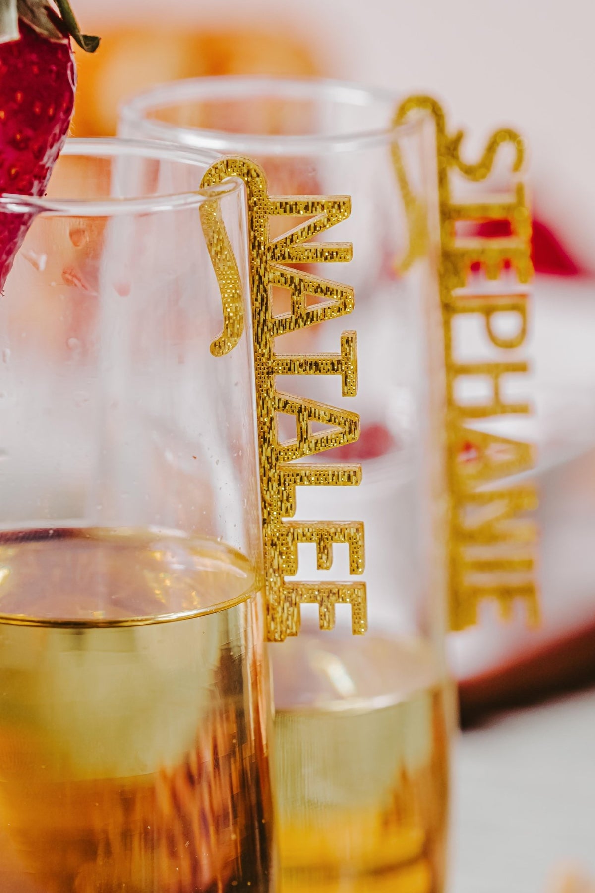 Two champagne glasses, each adorned with a Personalized Wine Glass Name Charm from Mirabilia Boutique, showcase custom acrylic drink tags—one “NATALEE,” the other “STEPHANIE”—with a strawberry on the rim for elegant, customized celebration decor.