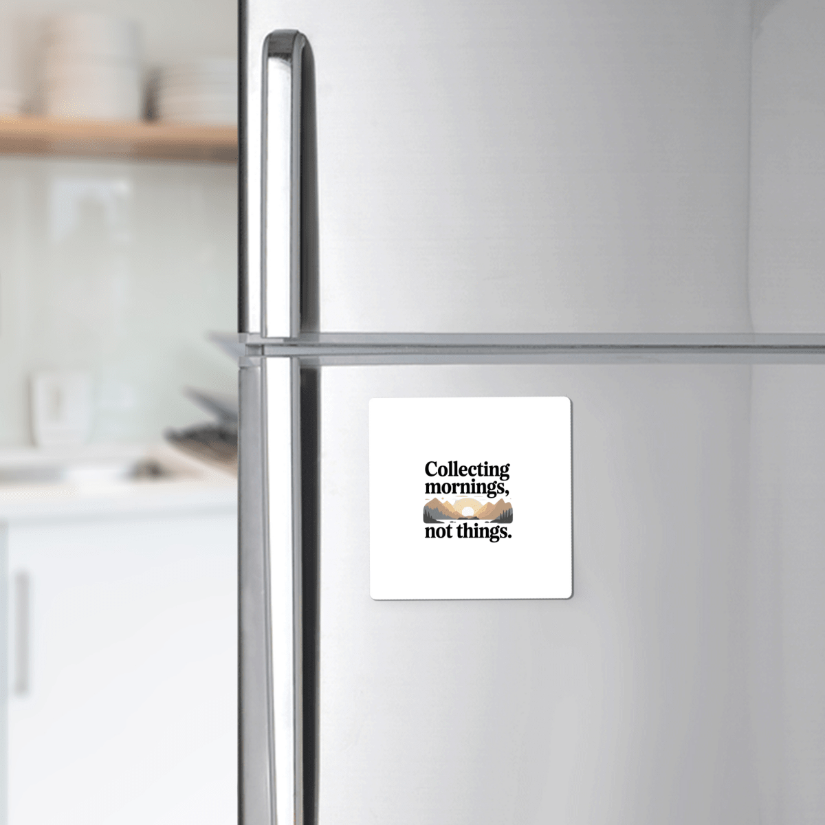 A Mirabilia Boutique "Collecting Mornings Not Things" Magnet sits on a stainless steel fridge, with stacked dishes and utensils in the blurred kitchen background—perfect adventure décor for outdoor enthusiasts.