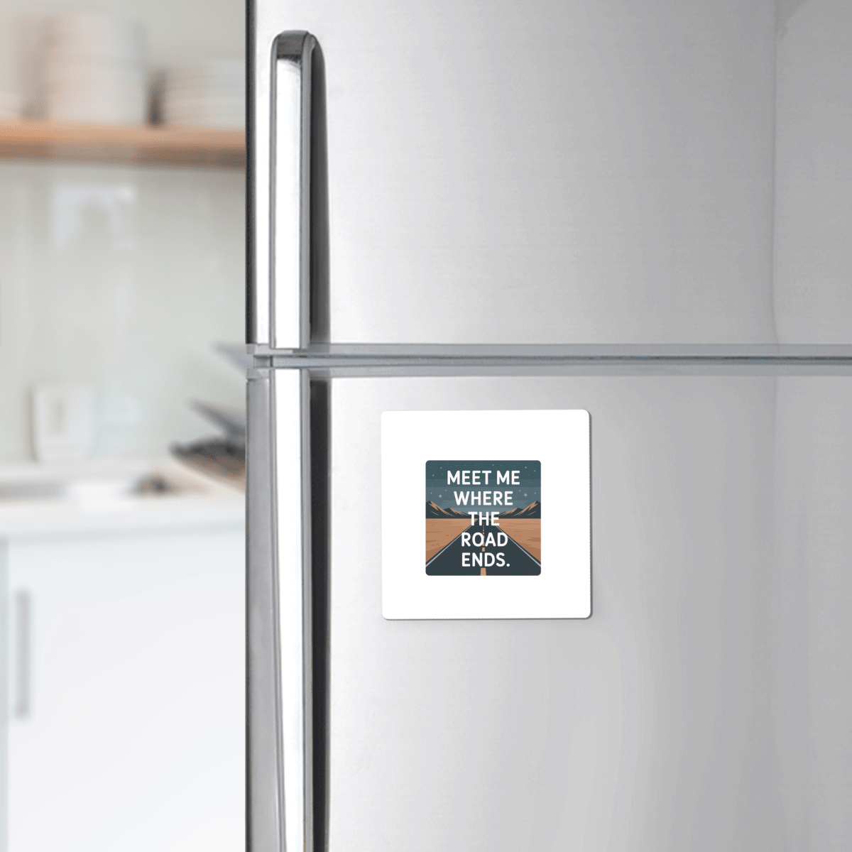 A silver fridge features the Mirabilia Boutique "Meet Me Where the Road Ends Magnet," a square design with a scenic road and bold text—ideal as camper decor or a unique road trip gift for any modern kitchen.