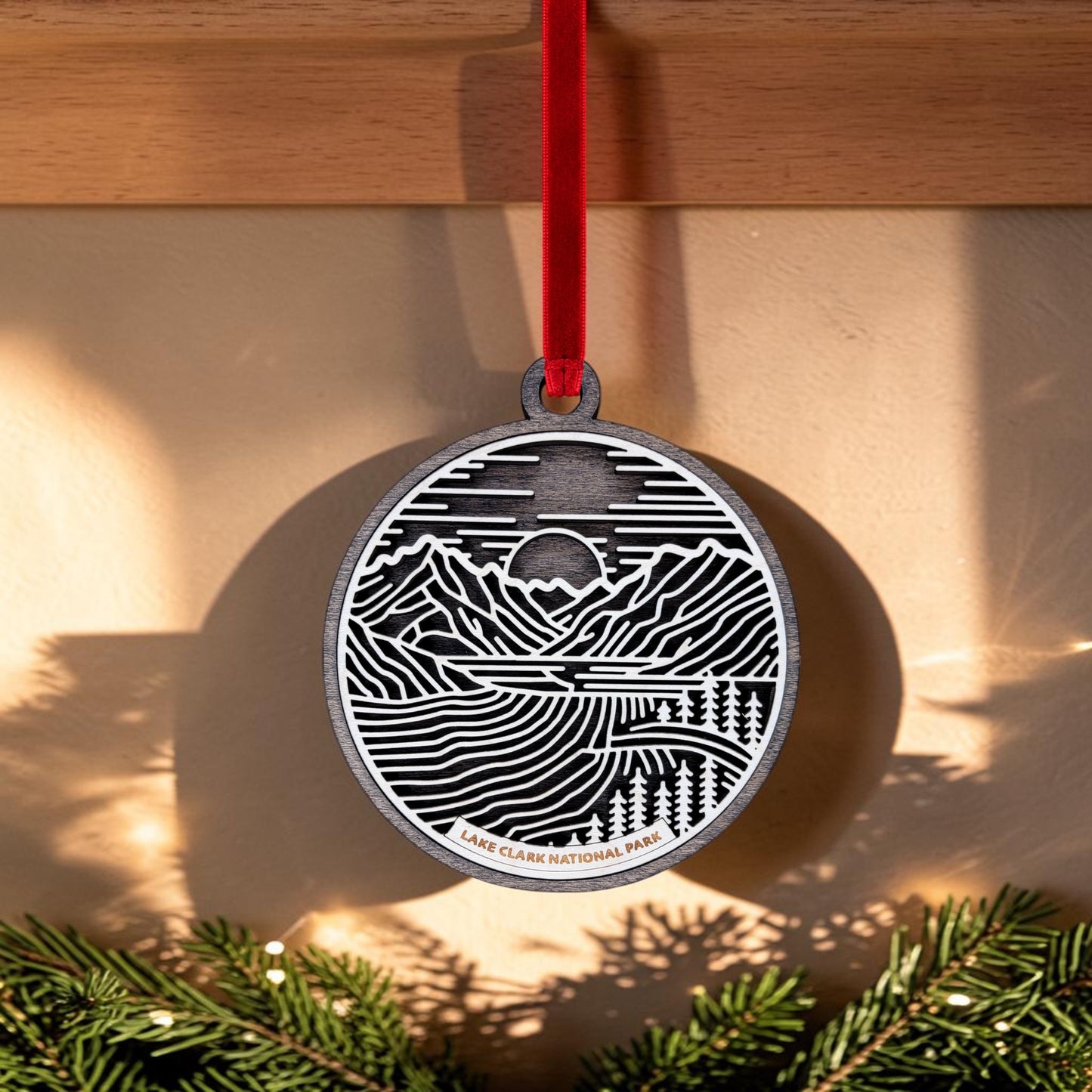 A handcrafted oval wooden ornament from Mirabilia Boutique, the "Lake Clark National Park Line Art Christmas Ornament," features a black and white engraved mountain scene labeled "Lake Clark National Park." Suspended from a red ribbon, this unique keepsake captures the rugged beauty of the Alaskan wilderness. Sunlight casts shadows on the wall while pine branches rest below.