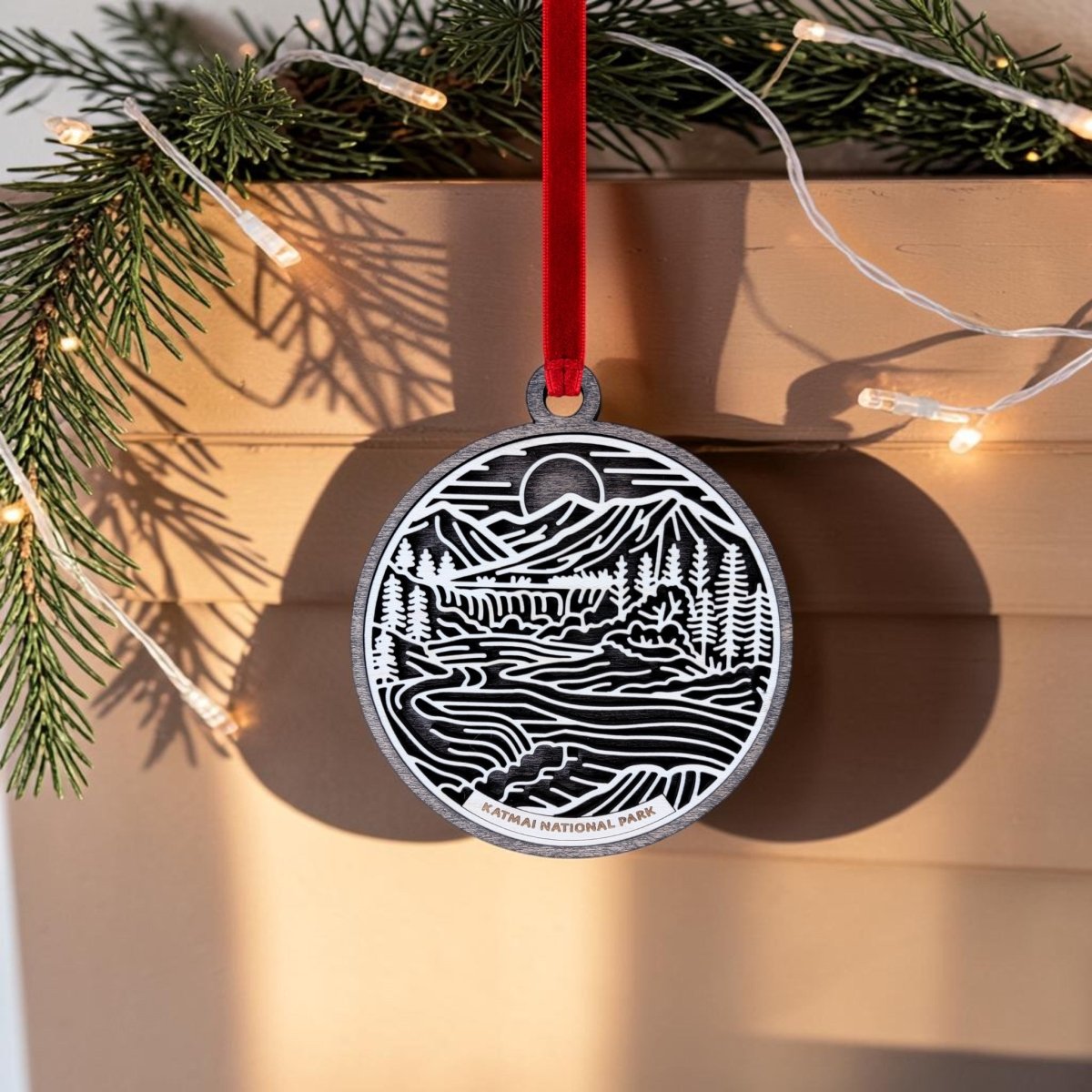 This handcrafted wooden ornament from Mirabilia Boutique features a line art scene of mountains, trees, and rivers labeled "Katmai National Park." It hangs by a red ribbon, with pine branches and string lights in the background.