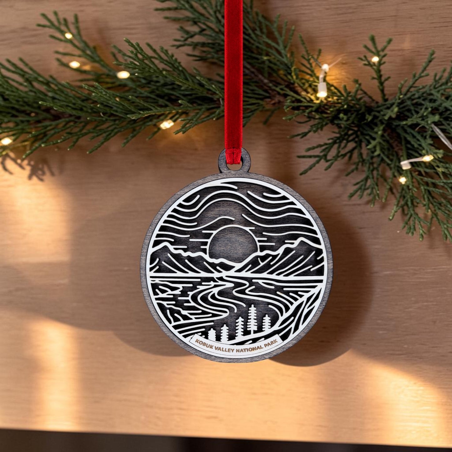 The Kobuk Valley National Park Line Art Christmas Ornament from Mirabilia Boutique features a handcrafted oval design with an intricate line art mountain scene, river, and trees, suspended from a red ribbon. Set against green pine branches adorned with small lights, this wooden ornament is inspired by the natural beauty of Kobuk Valley National Park and makes a delightful Alaska keepsake.