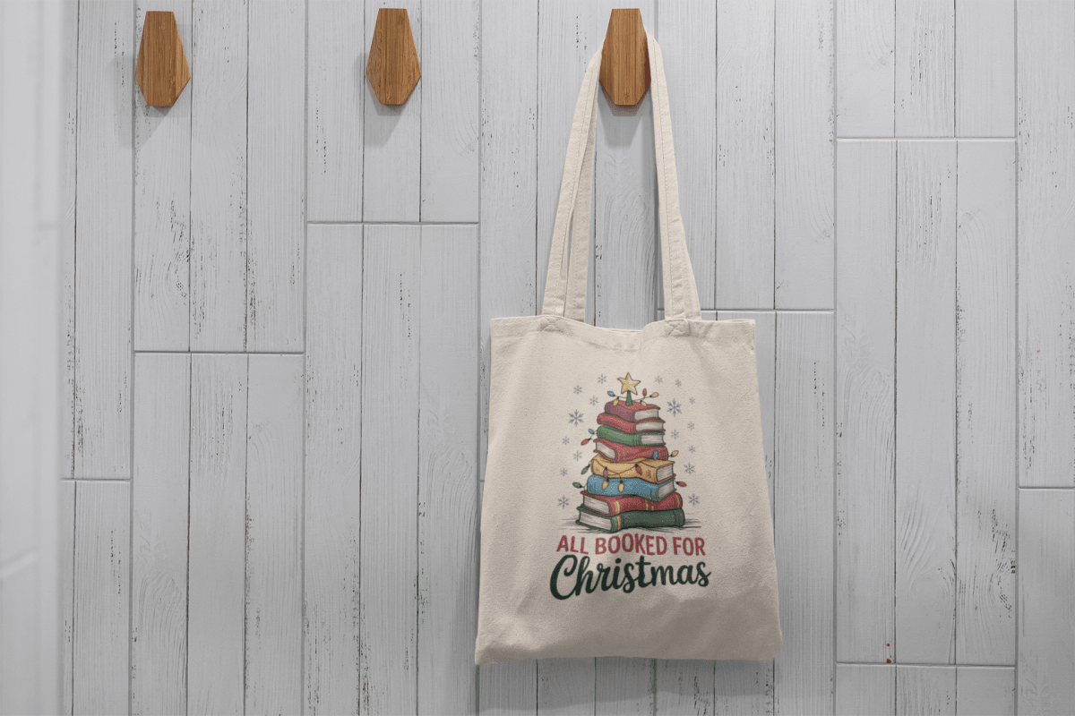 The Mirabilia Boutique "All Booked for Christmas Book Stack Tote Bag" hangs on a wooden hook against white tiles, featuring a festive stack of books and text—ideal as a holiday gift or reading bag for book lovers.
