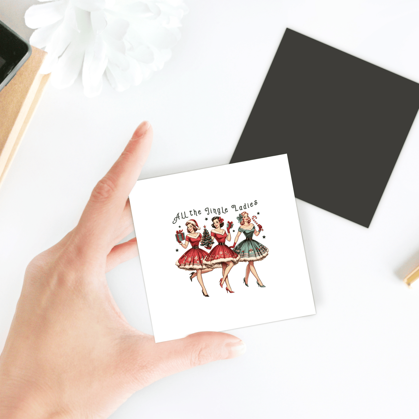 A hand holds the Mirabilia Boutique "All the Jingle Ladies Magnet"—a square, retro Christmas decor piece with three women in festive dresses and playful text, set against a white background. Other items are partially visible on the table.