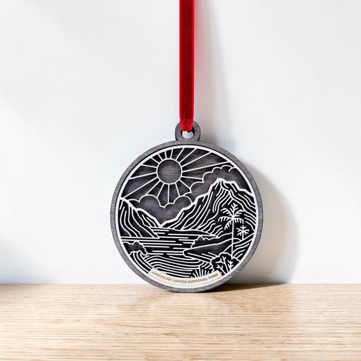 The American Samoa National Park Line Art Ornament by Mirabilia Boutique features a round, engraved design showcasing a scenic mountain, river, and sun inspired by the park’s natural beauty. Hanging from a red ribbon against a white wall and resting above a wooden surface, this handcrafted ornament beautifully captures the spirit of adventure and wonder.
