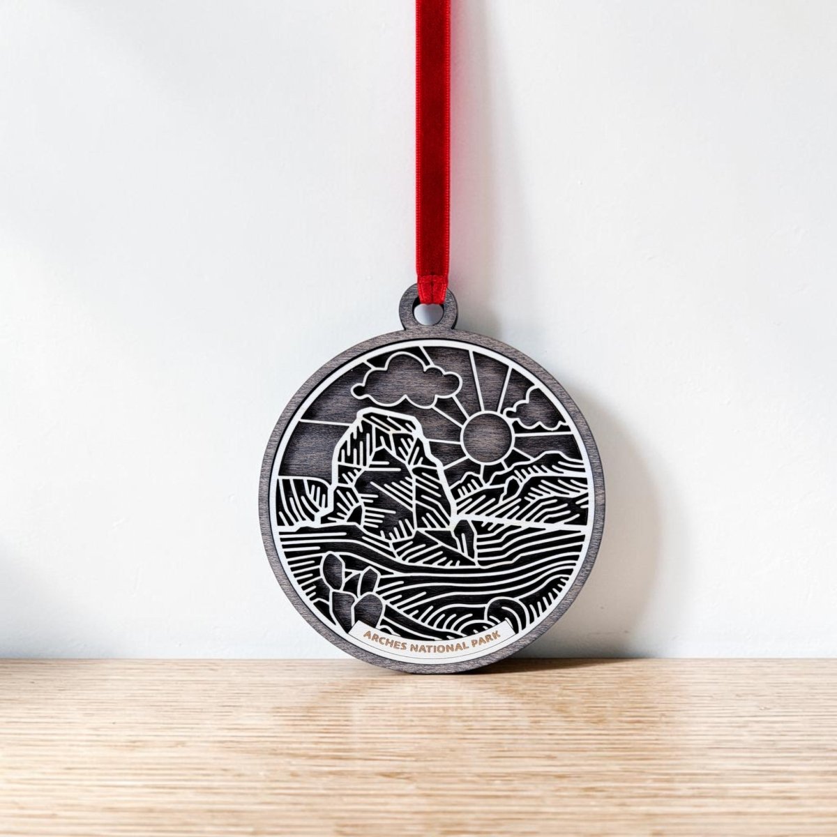 The Mirabilia Boutique Arches National Park Line Art Ornament is a round silver keepsake adorned with a red ribbon. It features an engraved landscape of iconic rock formations, drifting clouds, and the sun in delicate line art. This handcrafted ornament rests against a white wall on a wooden surface.