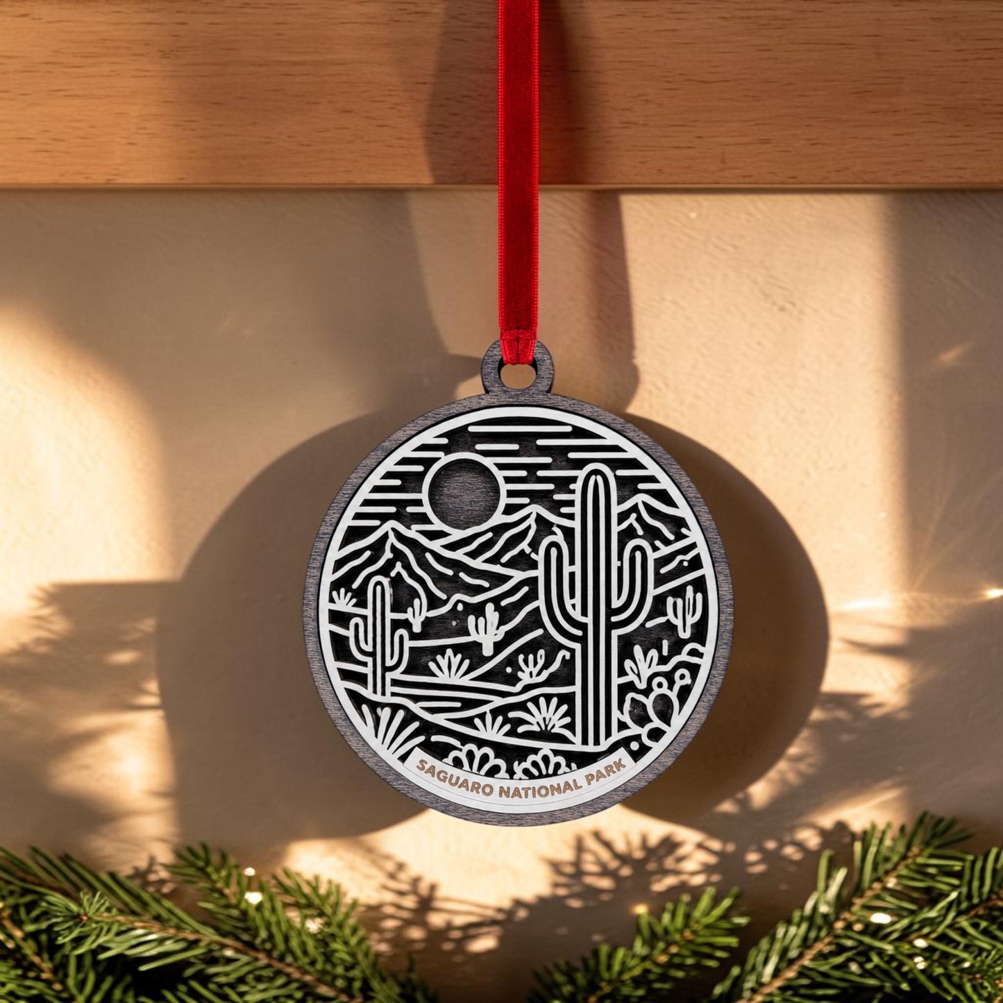 The Saguaro National Park Line Art Christmas Ornament by Mirabilia Boutique features a stunning desert landscape and cacti design. Hanging from a red ribbon against a wall, with sunlight and fir branches below, this wooden ornament is the perfect keepsake for any nature lover or fan of desert landscapes.