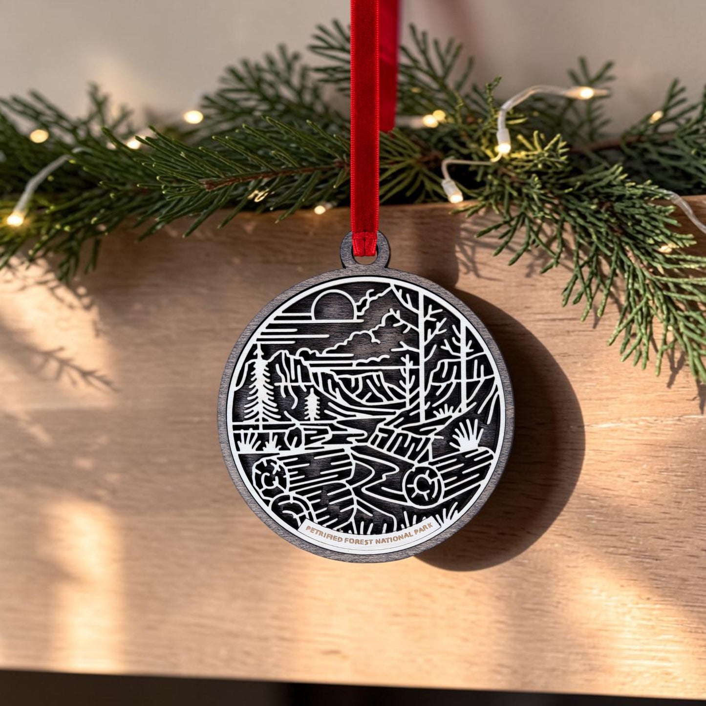 The Petrified Forest National Park Line Art Christmas Ornament by Mirabilia Boutique features an oval wooden design displaying a nature scene with trees, mountains, and a river set against a desert landscape. It hangs from a red ribbon in front of green pine branches and twinkling lights on a wooden surface.