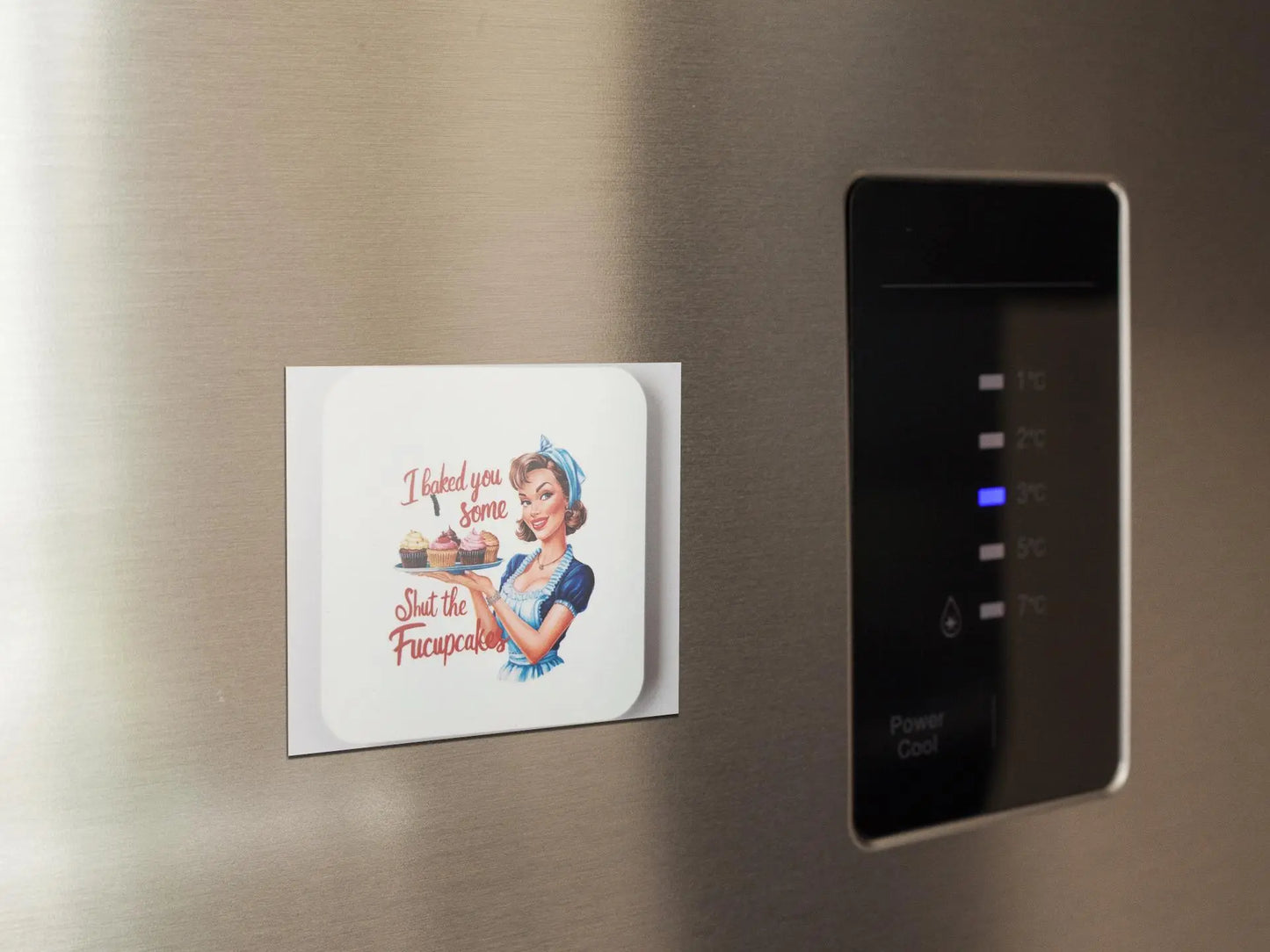 An "I Baked You Some Shut The Fucupcakes" magnet by Mirabilia Boutique featuring a retro-style illustration of a woman holding a pie. The text humorously reads, "I baked you some Shut the Frucupcakes." This funny fridge magnet is affixed to a stainless steel fridge door adjacent to a digital control panel displaying temperature settings.