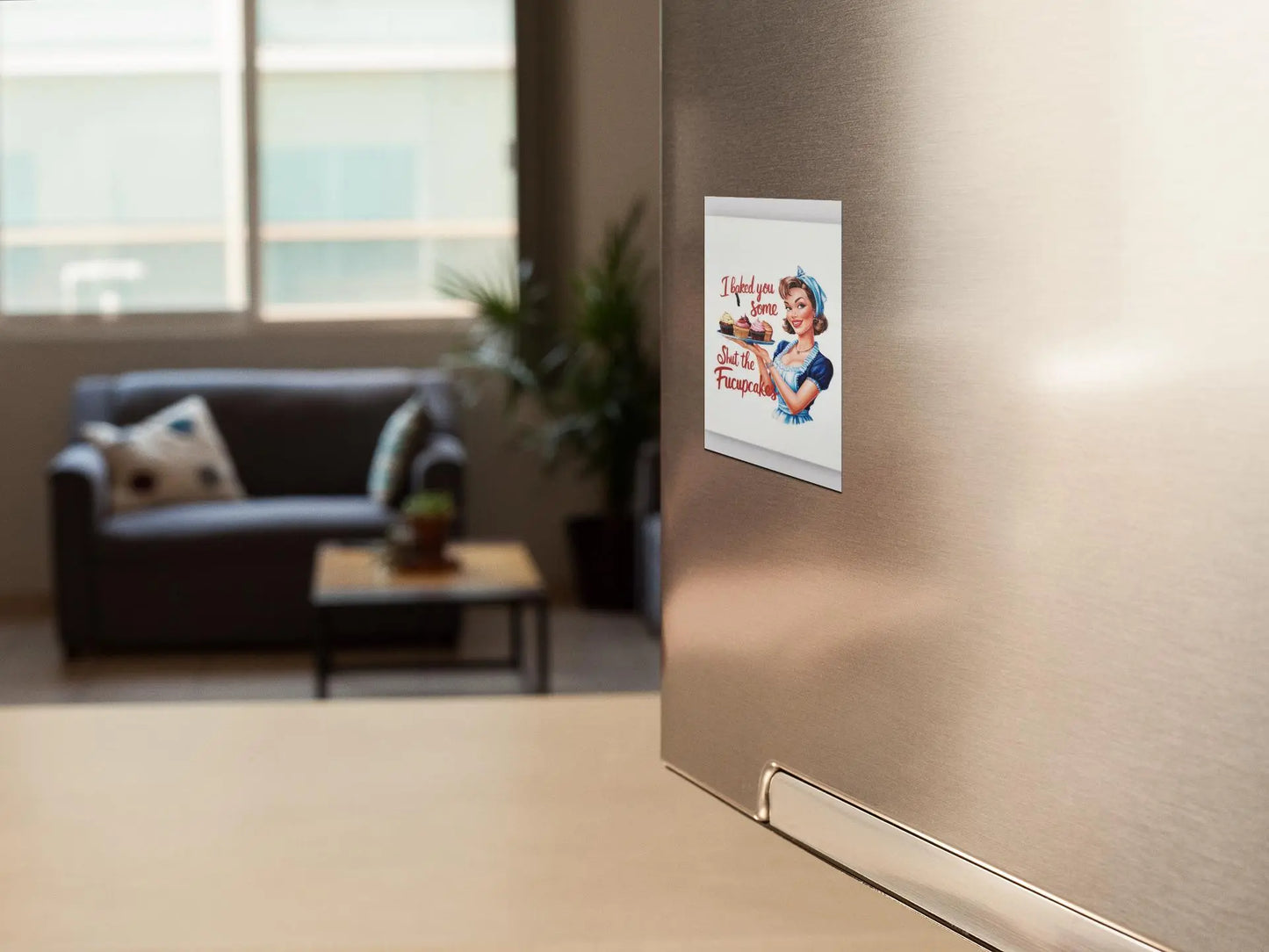 A kitchen scene shows a refrigerator adorned with a small, funny fridge magnet from Mirabilia Boutique that reads "I Baked You Some Shut The Fucupcakes." In the background, there's a cozy living area with a couch, pillows, and a plant—an ideal setting for such a unique gift.