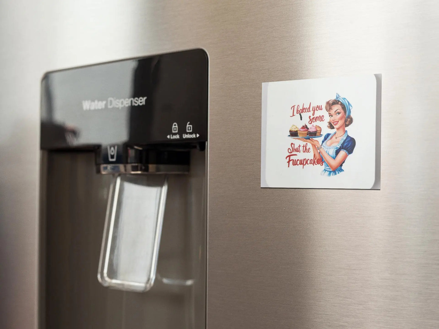 A refrigerator door with a water dispenser on the left side and a funny Mirabilia Boutique magnet on the right featuring an illustration of a retro housewife holding a tray of cupcakes. The magnet text reads, "I baked you some Shut the Fucupcakes!" Perfect as a unique gift for those who appreciate humor, this product is titled "I Baked You Some Shut The Fucupcakes".