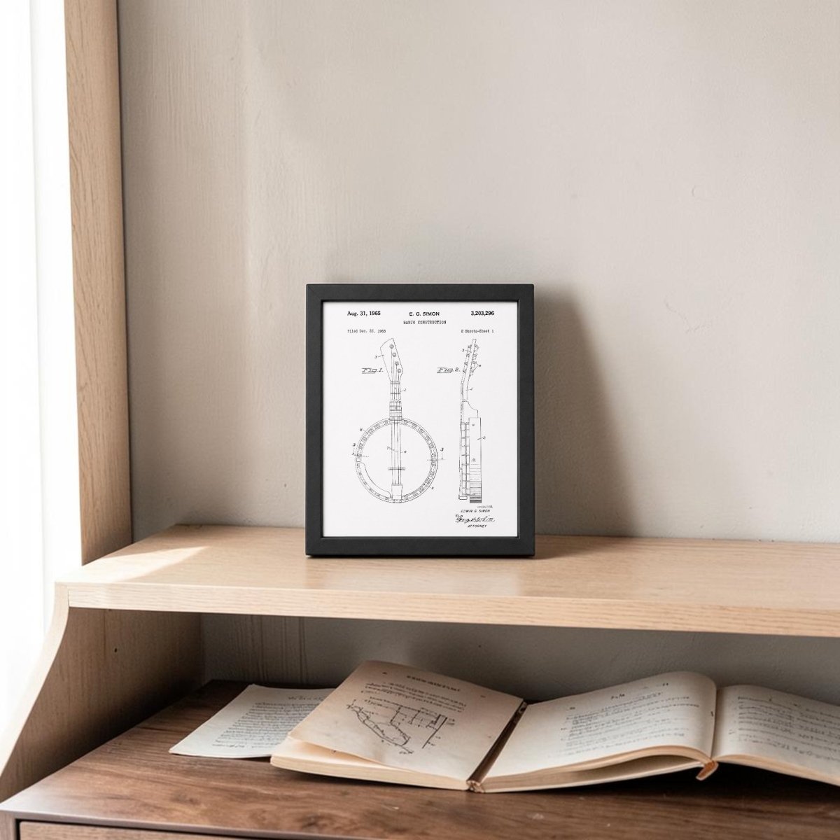 The Banjo Patent Print 8x10 Wall Art by Mirabilia Boutique is displayed in a frame on a wooden shelf against a light wall. Below, an open book with diagrams and another book sit, all highlighted by natural light from the left for vintage charm.