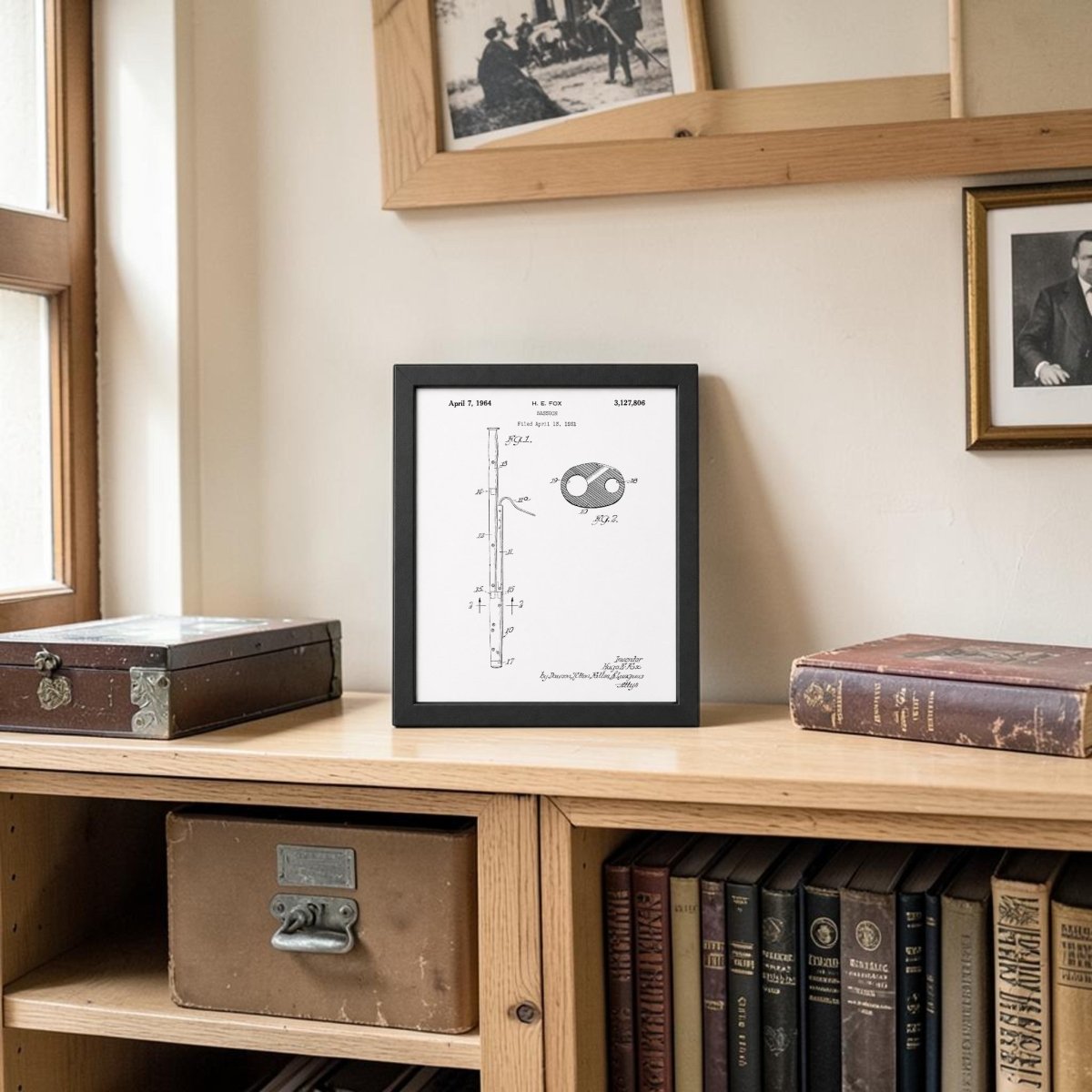 The Mirabilia Boutique Bassoon Patent Print on Metal Wall Art is displayed on a shelf beside books and boxes, with framed photos above. Sunlight from a nearby window highlights this distinctive bassoon decor piece.