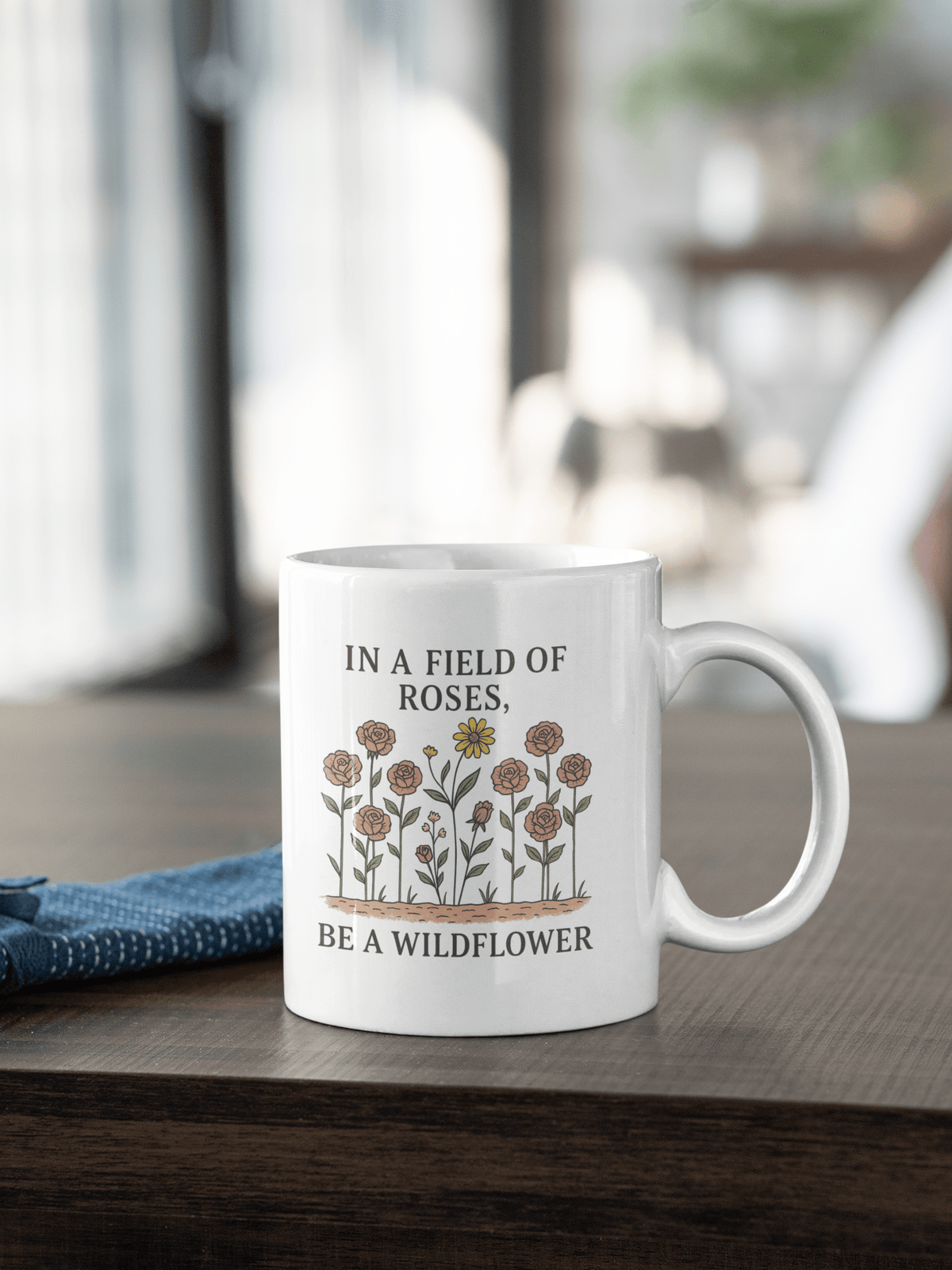 The Be a Wildflower Mug by Mirabilia Boutique is a white ceramic coffee cup with a motivational floral design and the quote "In a field of roses, be a wildflower," perfect for cozy indoor moments.