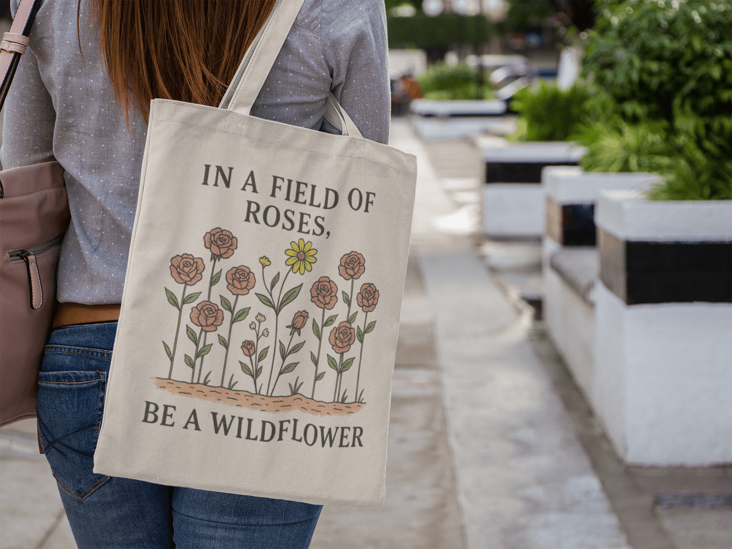 A woman walks outdoors with the Mirabilia Boutique "Be a Wildflower Tote Bag," an eco-friendly cotton canvas tote featuring rose and yellow wildflower illustrations and the quote, "IN A FIELD OF ROSES, BE A WILDFLOWER.