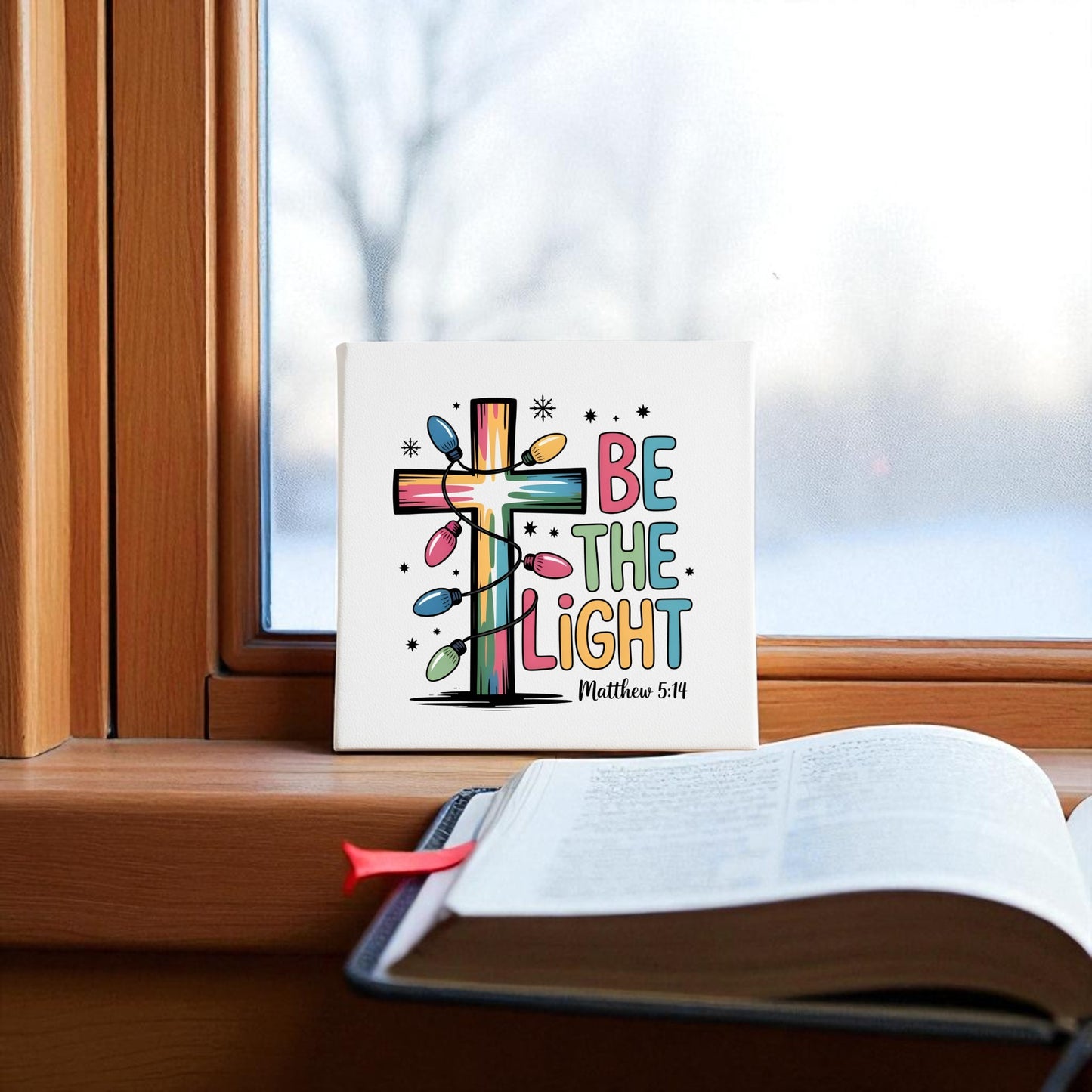 The "Be the Light | Faith-Inspired Christmas Decor | Matthew 5:14" sign by Mirabilia Boutique sits on a windowsill beside an open Bible, with snowy trees outside, creating a festive, faith-inspired holiday scene.