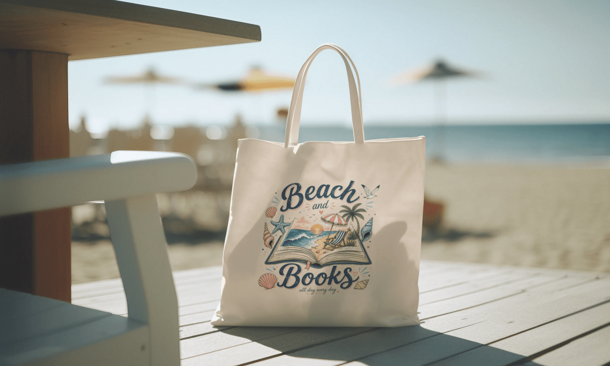 The Mirabilia Boutique "Beach and Books Canvas Tote Bag" features a beach scene illustration and text, shown on a wooden table by the ocean with sand and sun umbrellas in the background.