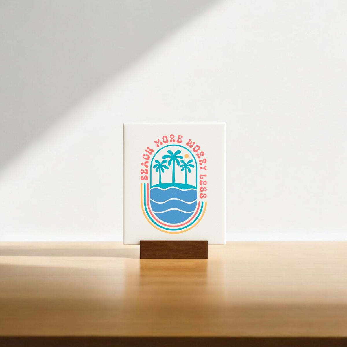 The Mirabilia Boutique "Beach More Worry Less Retro Summer Sign" features palm trees over waves in colorful retro style on a wooden stand—ideal as beach decor or a fun summer gift. Sunlight casts a soft shadow for added charm.
