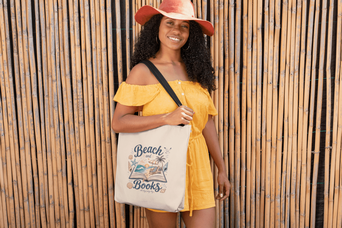A smiling woman in a yellow romper and orange hat stands by a bamboo wall, holding the Beach and Books Canvas Tote Bag from Mirabilia Boutique, featuring "Beach and Books" with an open book, palm trees, and sea illustration.