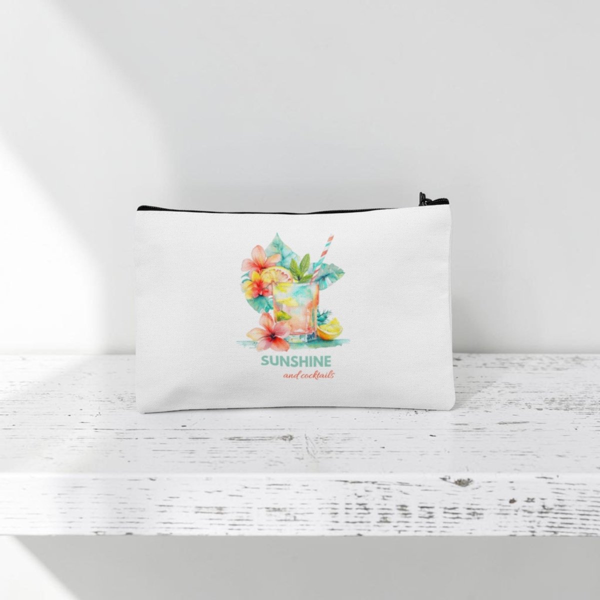 The Mirabilia Boutique Sunshine and Cocktails Makeup Bag—a white zippered travel pouch featuring a vibrant tropical drink, colorful flowers, and the phrase "SUNSHINE and cocktails" in decorative text—rests on a white wooden surface with a white background.