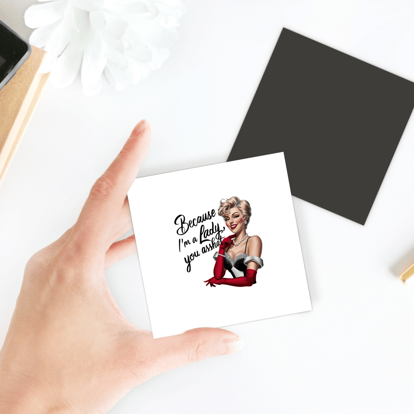 A hand holds Mirabilia Boutique’s "Because I’m a Lady" sarcastic retro UV-printed magnet, featuring a glamorous woman and the phrase, "Because I'm a lady, you a**hole"—a funny gift for women or friends. Black square and white flower included.