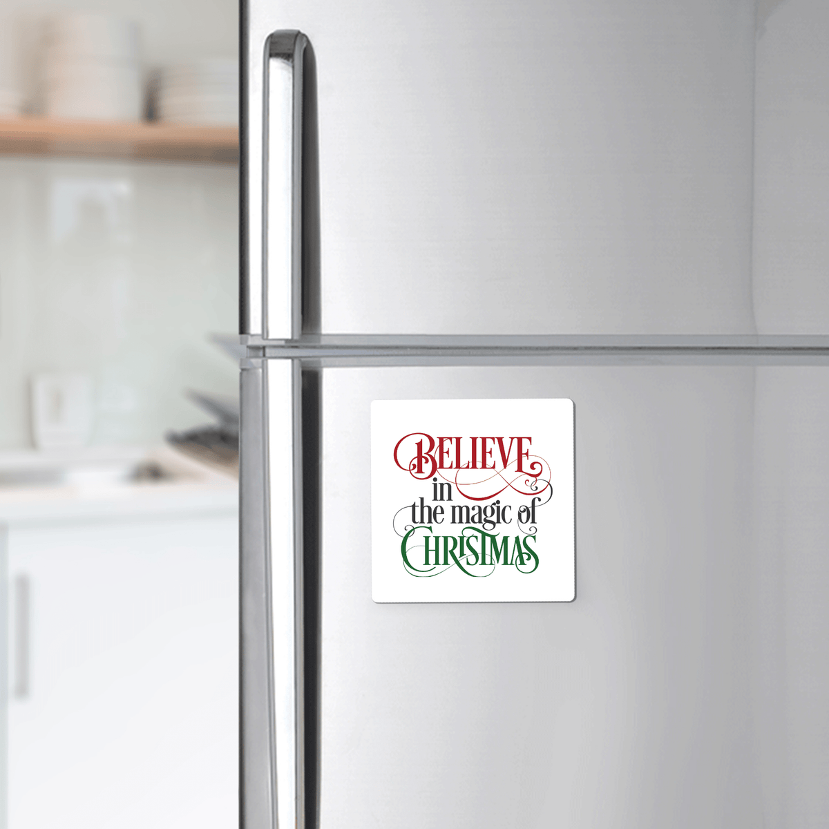 The Believe in the Magic of Christmas Magnet by Mirabilia Boutique features festive red and green lettering—perfect for holiday gifting or as a stocking stuffer. Blurred kitchen shelves and utensils appear in the background.