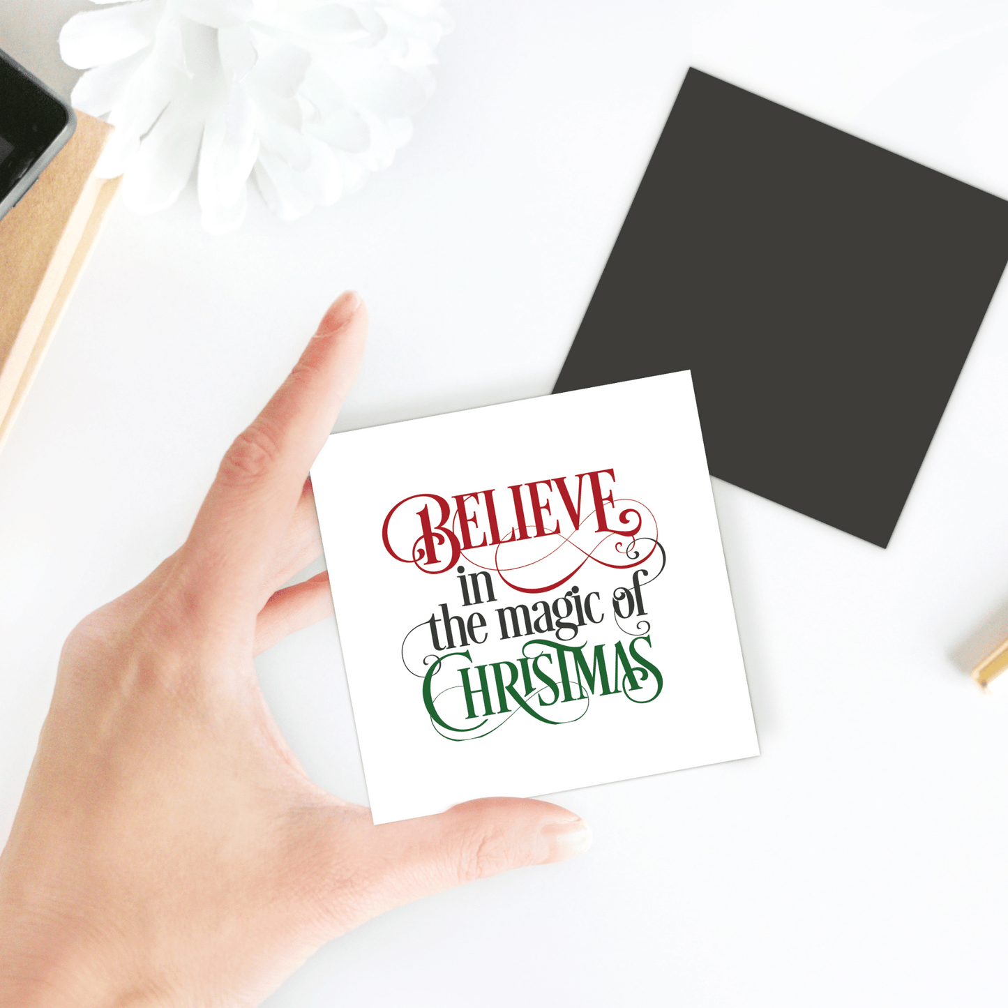 A hand displays the Mirabilia Boutique "Believe in the Magic of Christmas" magnet—festive fridge decor, ideal as a holiday gift or stocking stuffer. A white flower, black square, and notebook sit on a white surface in the background.