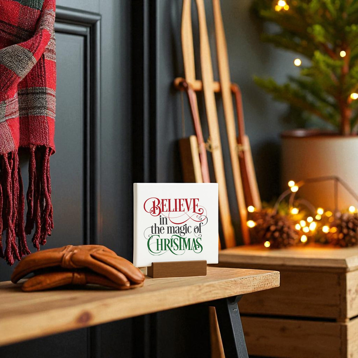 A cozy holiday scene features the Mirabilia Boutique "Believe in the Magic of Christmas" 4x4 mini sign on a wooden table, surrounded by gloves, pinecones, festive decor, a plaid blanket, sleds, and a glowing Christmas tree in the background.
