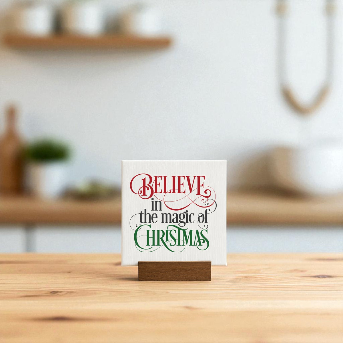 The Mirabilia Boutique "Believe in the Magic of Christmas" Mini Sign (4x4) sits on a table, displaying its festive red and green script. A softly blurred kitchen with holiday decor provides a cozy seasonal backdrop.