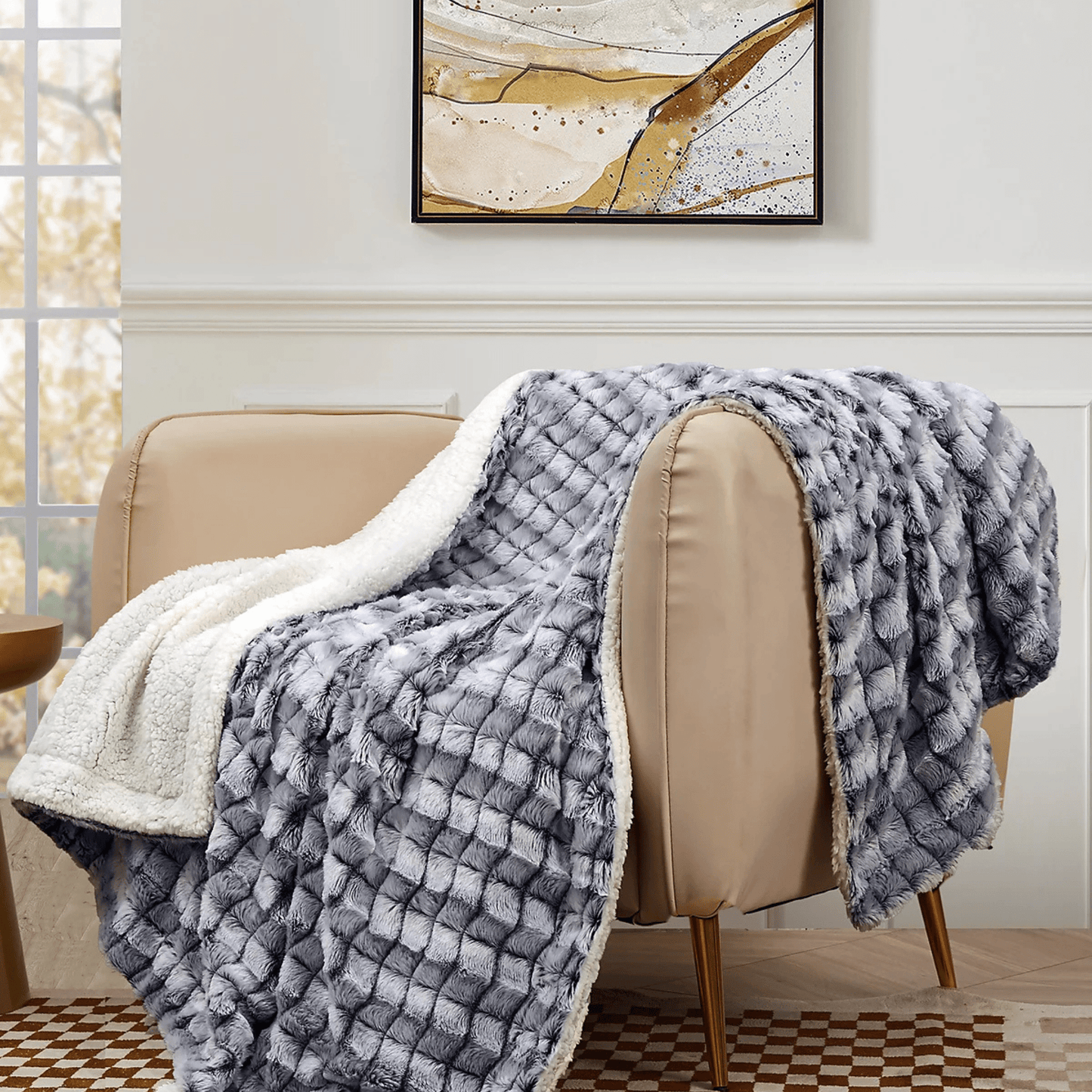 The Mirabilia Boutique Custom Fall Throw Blanket, featuring a soft gray and white design with plush white lining, is draped over a beige armchair in a bright, cozy room beneath an abstract painting.