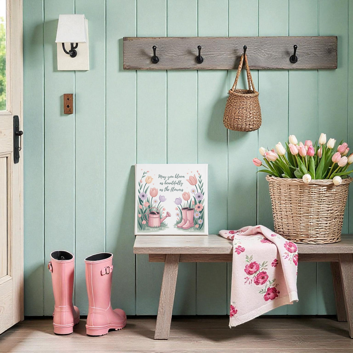 A cozy entryway with mint green walls, a wooden bench, pink rain boots, pink tulips in a basket, and spring decor—a Mirabilia Boutique "Bloom Like the Flowers Canvas Art – Spring Garden Quote" hanging above in a wicker bag on wall hooks.