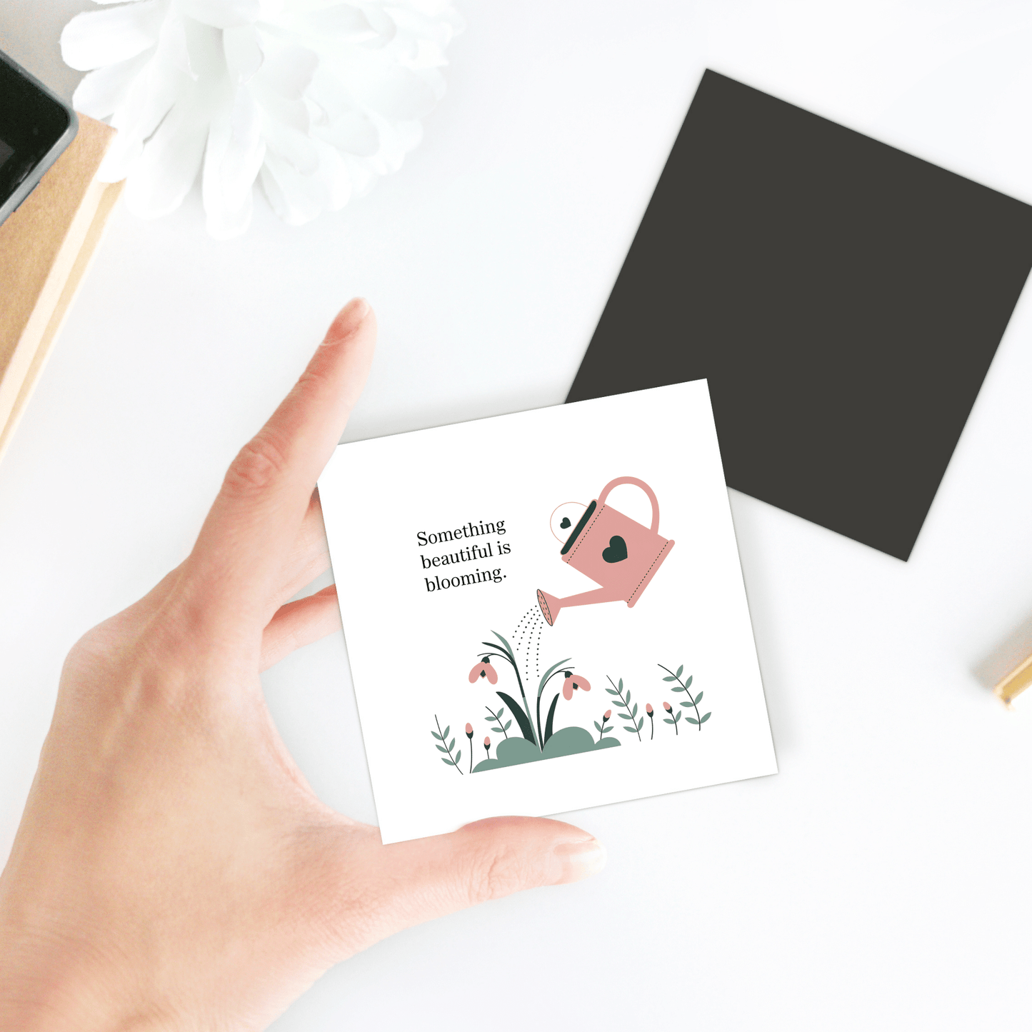 A hand holds Mirabilia Boutique's "Something Beautiful Is Blooming" magnet, featuring a pink watering can and flowers. On the white table are a black square and a white flower—perfect growth-inspired decor for self-care gifts.