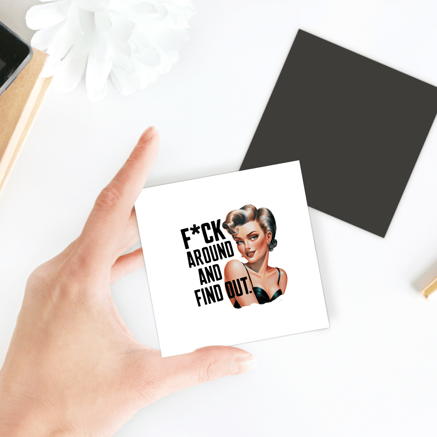 A hand holds a Mirabilia Boutique "F*ck Around and Find Out" 3x3 retro UV printed magnet with a bold pinup design—perfect as a sassy gift for friends, coworkers, or office desks. Black square and white flower nearby on the table.