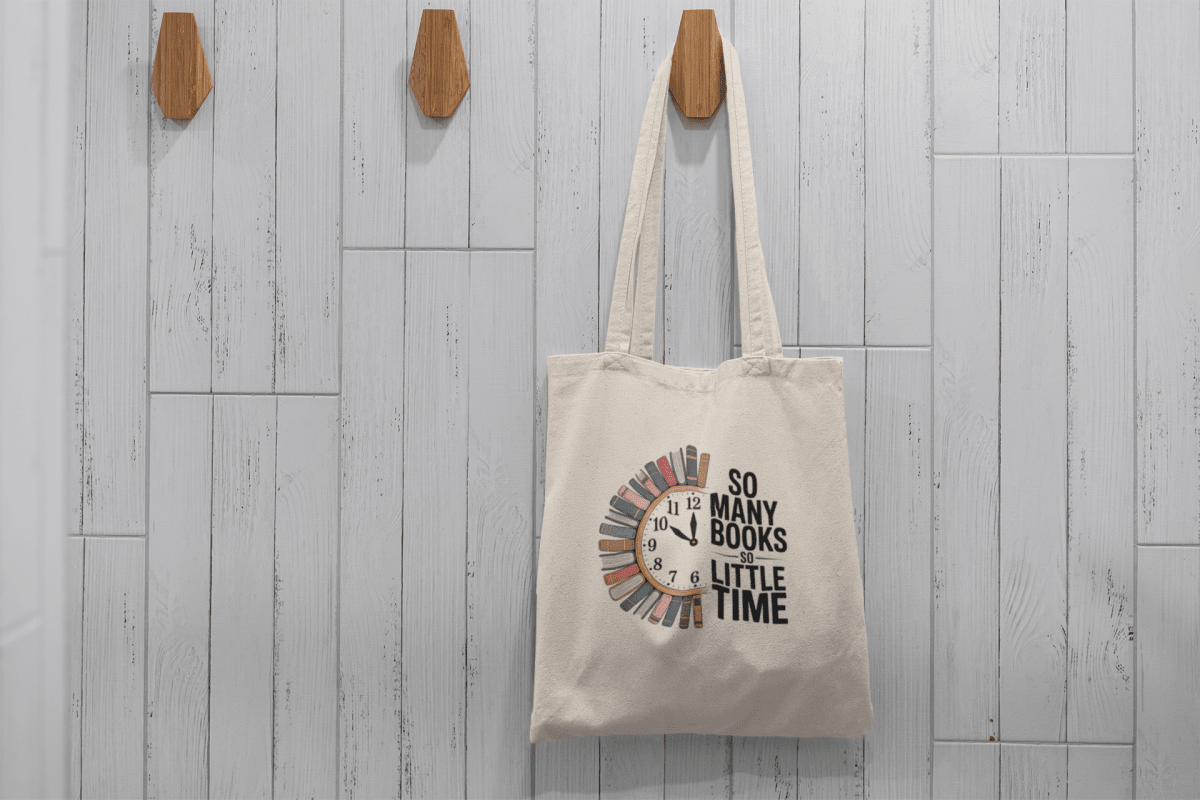 The So Many Books Canvas Tote Bag by Mirabilia Boutique, featuring a clock-shaped stack of books and "So many books, so little time," hangs on a wooden hook—perfect as a book lovers gift or stylish library tote.