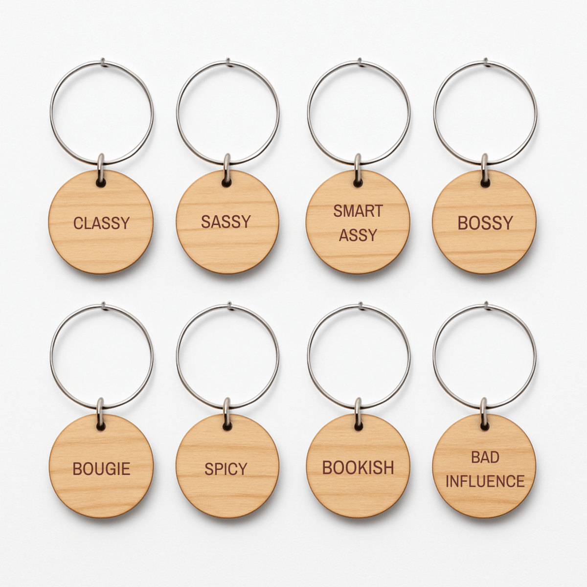 Eight round wooden keychains with metal rings from Mirabilia Boutique’s “Bridal Party Personality Wine Charms | Set of 8” are arranged in two rows. Each charm showcases a distinct engraved word—“CLASSY,” “SASSY,” “SMART ASSY,” “BOSSY,” “BOUGIE,” “SPICY,” “BOOKISH,” and “BAD INFLUENCE.” Perfect as unique bridal party wine charms or thoughtful gifts, these wooden accents add a playful touch to any celebration.