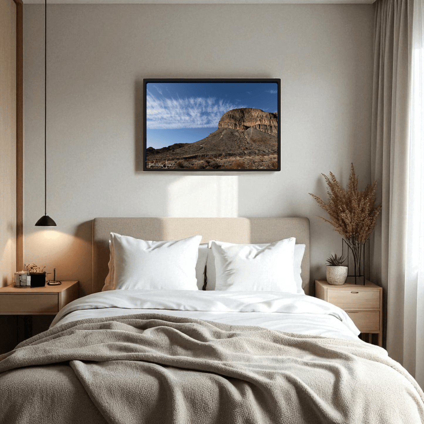 A cozy bedroom with light bedding, wooden side tables, and a hanging lamp features the "Burro Mesa Pouroff Print | Big Bend Framed Wall Art" by Mirabilia Boutique above the bed. Dried plants decorate one side table.