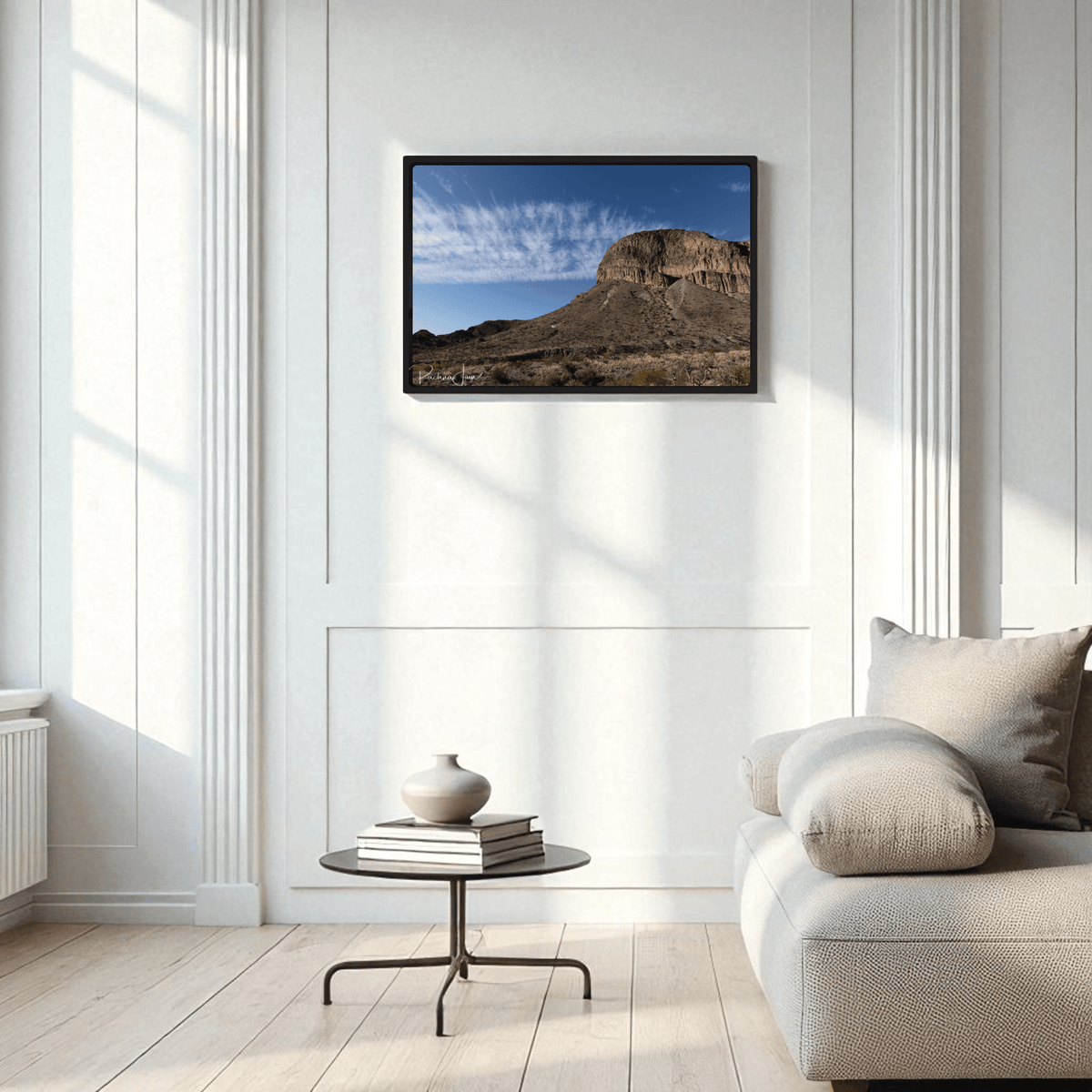 A minimalist living room with a light gray sofa, round black coffee table, and vase features the Burro Mesa Pouroff Print | Big Bend Framed Wall Art by Mirabilia Boutique on a white paneled wall.