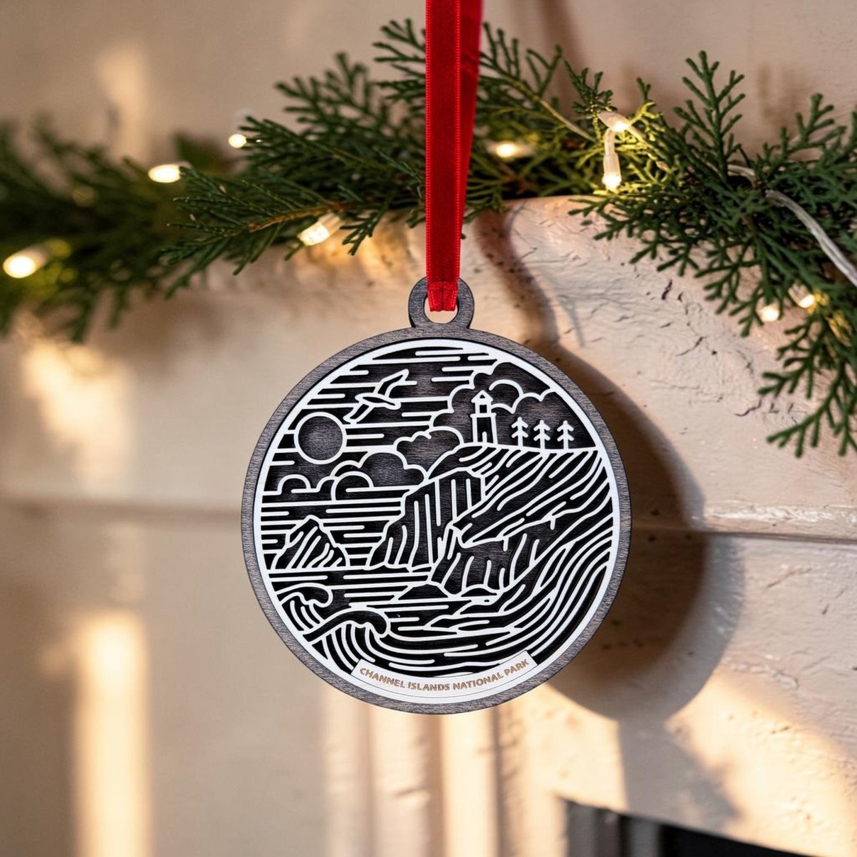 Certainly! Here's a rewritten version of the sentence using your product data: The Channel Islands National Park Christmas Ornament by Mirabilia Boutique is an oval wooden decoration showcasing black and white line art of the park's landscape, elegantly suspended from a vibrant red ribbon. In the background, green pine branches twinkle with string lights.