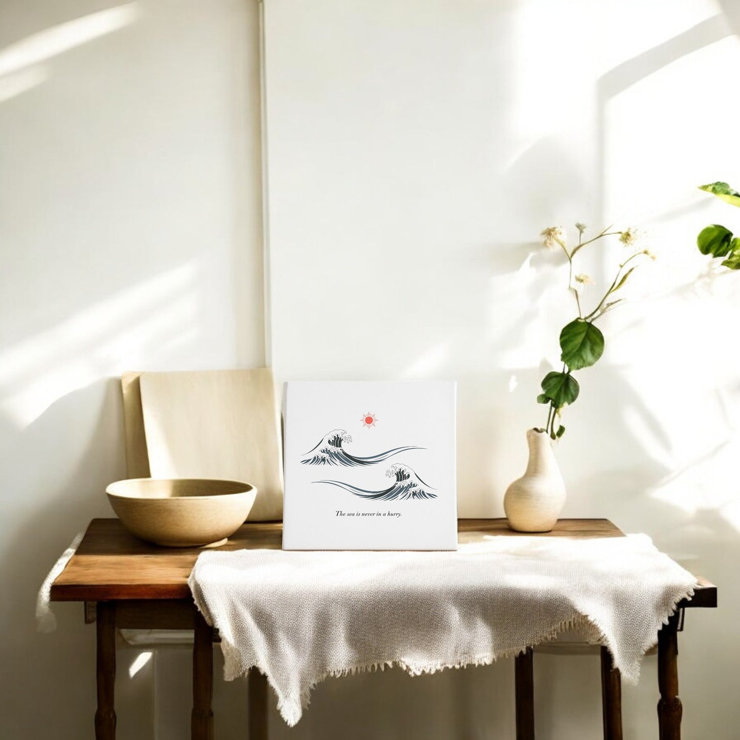 A sunlit table draped in white displays a ceramic bowl, leafy vase, and "The Sea Is Never in a Hurry Canvas – Serene Ocean Calm" by Mirabilia Boutique. Light streams in, creating a calm, airy space with minimalist ocean art accents.
