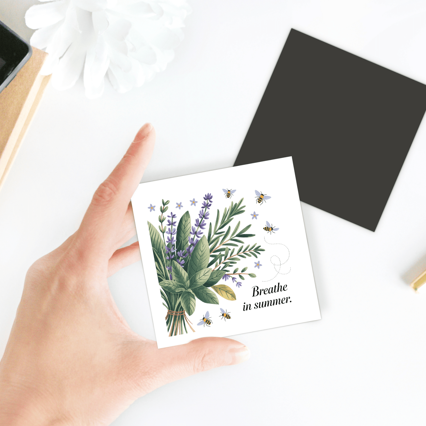 A hand holds the Mirabilia Boutique "Breathe in Summer Magnet – Botanical Calm & Gentle Bees," a square magnet card featuring herb, flower, and bee illustrations. A black envelope and white flower are on the surface in the background.